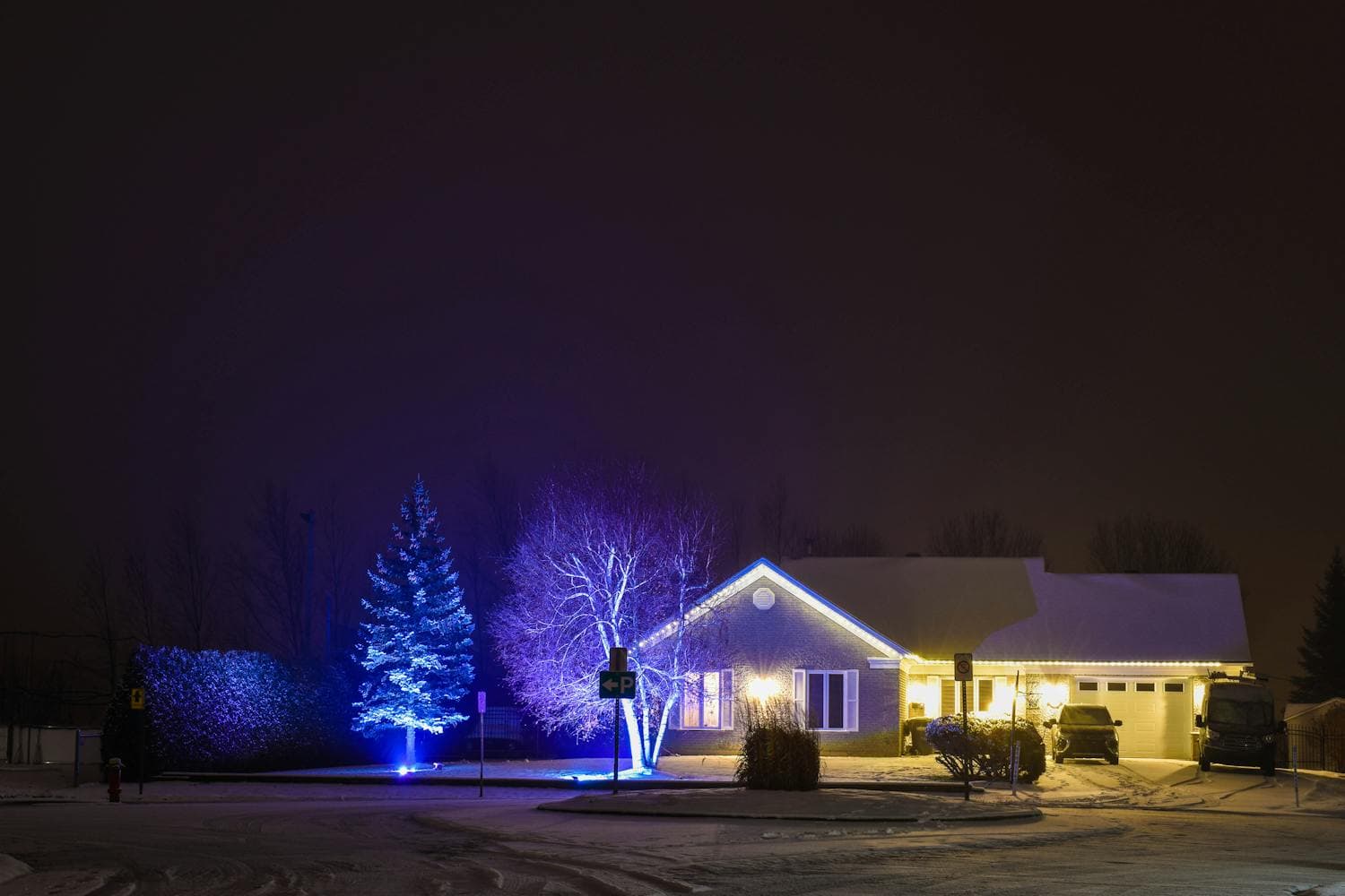 A single-story home at night with warm-white Christmas lights running continuously along the entire front roofline, snow on the lawn, and softly lit shrubs framing the path to the front door
