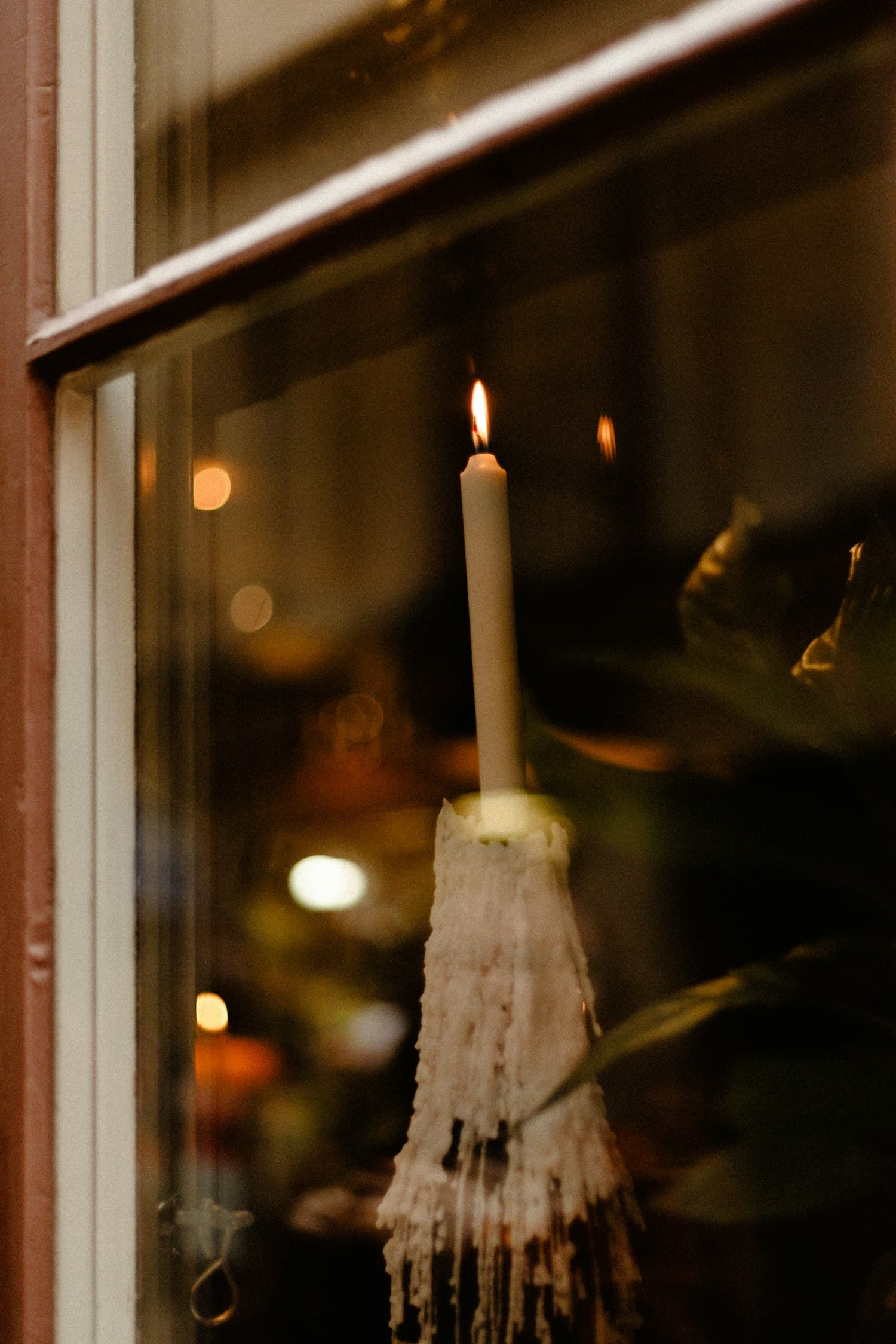 A single warm-white taper candle glowing in the lower pane of a wooden-framed window at dusk, photographed from outside through the glass