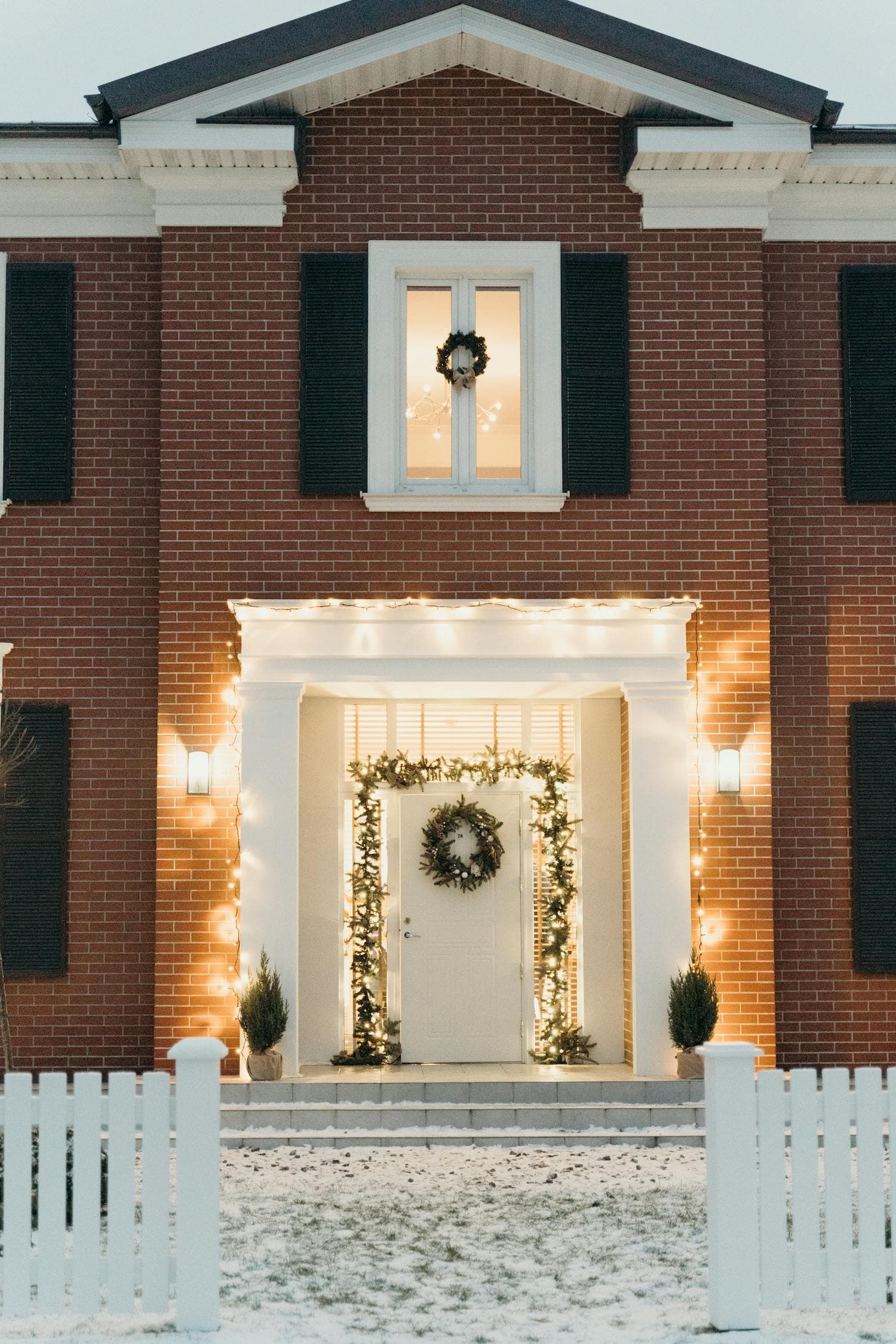 A brick colonial home in winter with a fresh evergreen wreath on the front door, warm-white light glowing from the windows, and snow visible on the front steps, the symmetric facade reading as one composed Christmas scene from the street