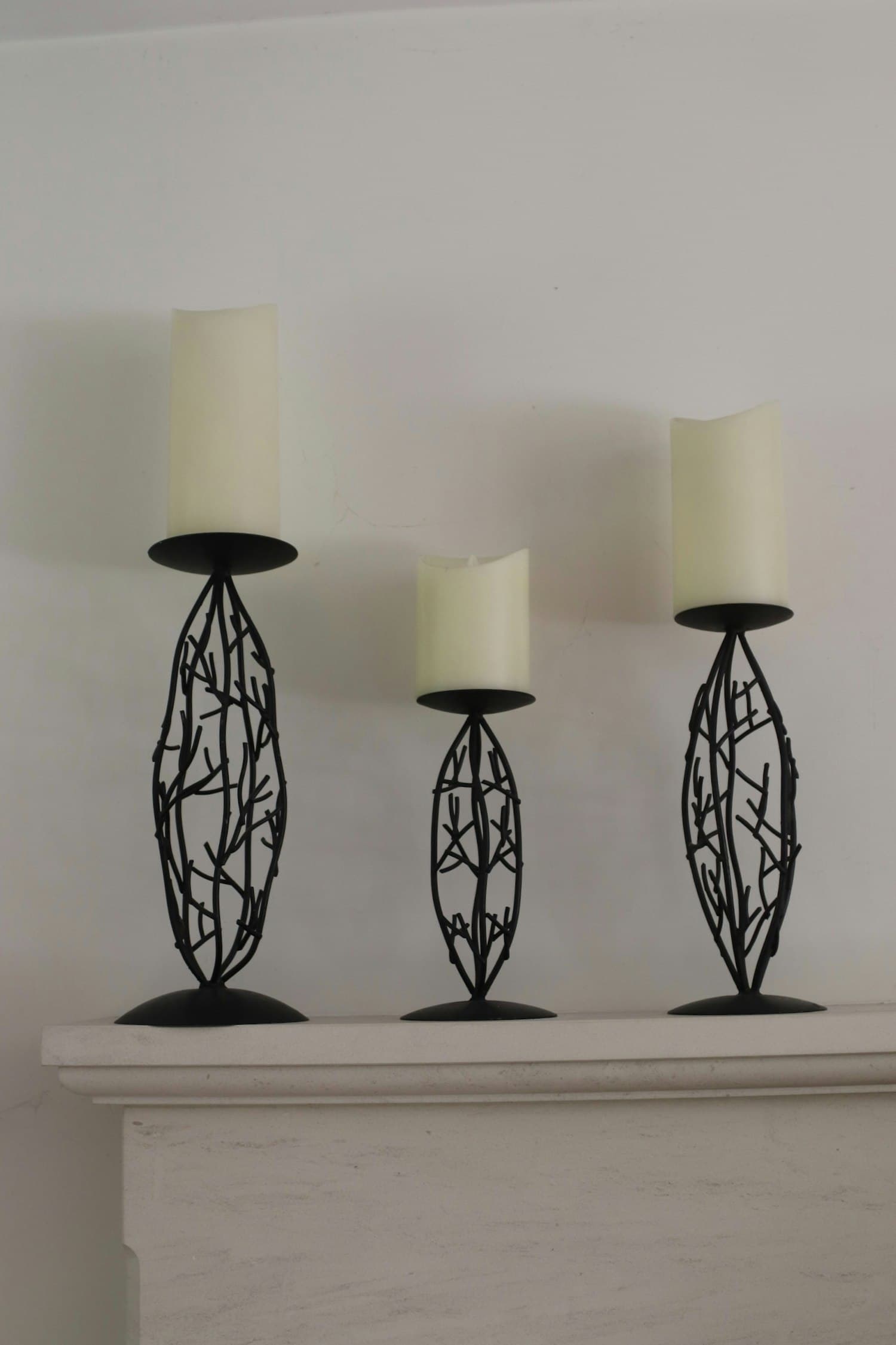 Three black wrought-iron candle holders with twig-motif bases on a clean white mantel, holding cream pillar candles in varied heights, against a soft white wall