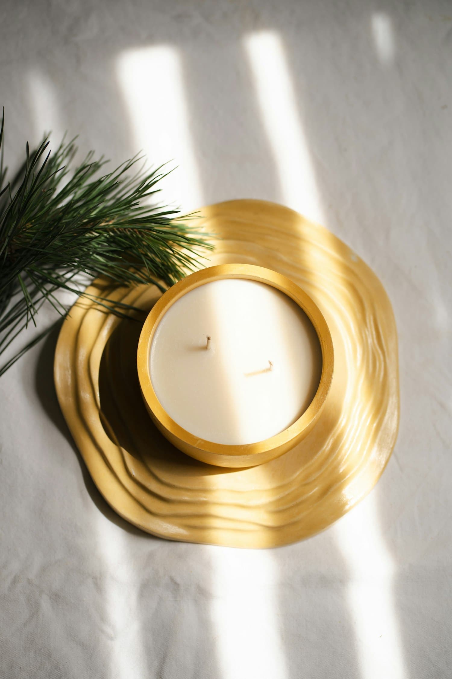A fresh pine sprig laid beside a cream pillar candle in a brass dish, on a linen surface lit by golden hour sunlight