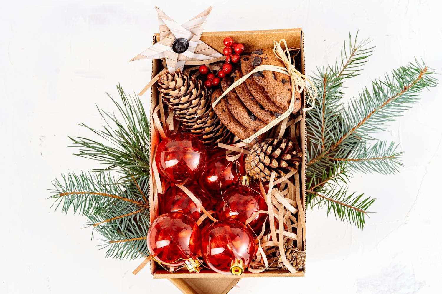 Overhead flat lay of a kraft paper box filled with red glass ornaments, pinecones, gingerbread cookies, raffia, and pine branches