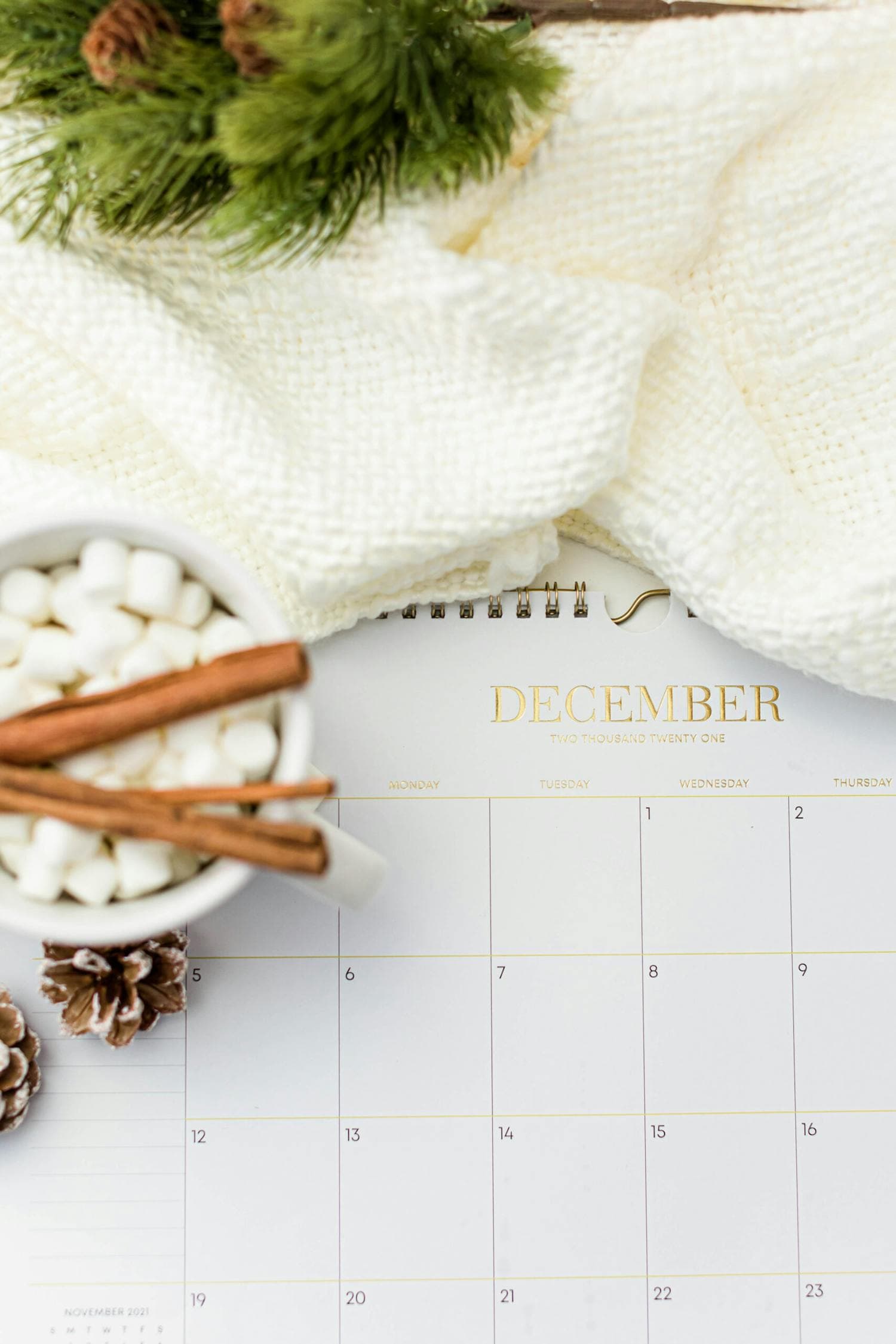 A flat lay of a December calendar surrounded by a steaming mug of hot chocolate with marshmallows, fresh pine sprigs, cinnamon sticks, and dried orange slices, the visual of a cozy planning session for the holiday season