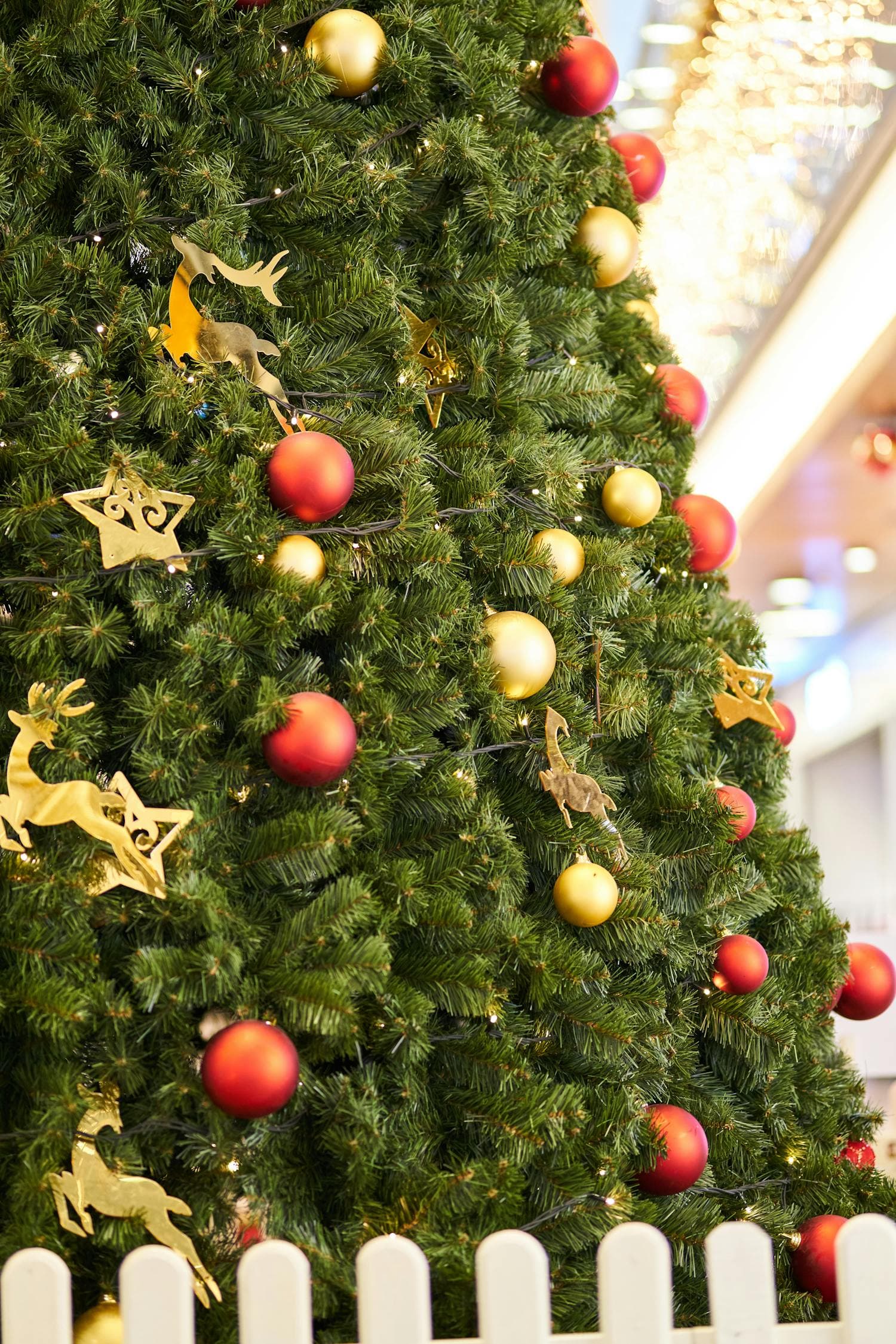 A fully decorated traditional Christmas tree with red velvet ribbon woven through the branches, deep red and gold glass ornaments layered at varying depths, and warm yellow string lights glowing against forest-green pine