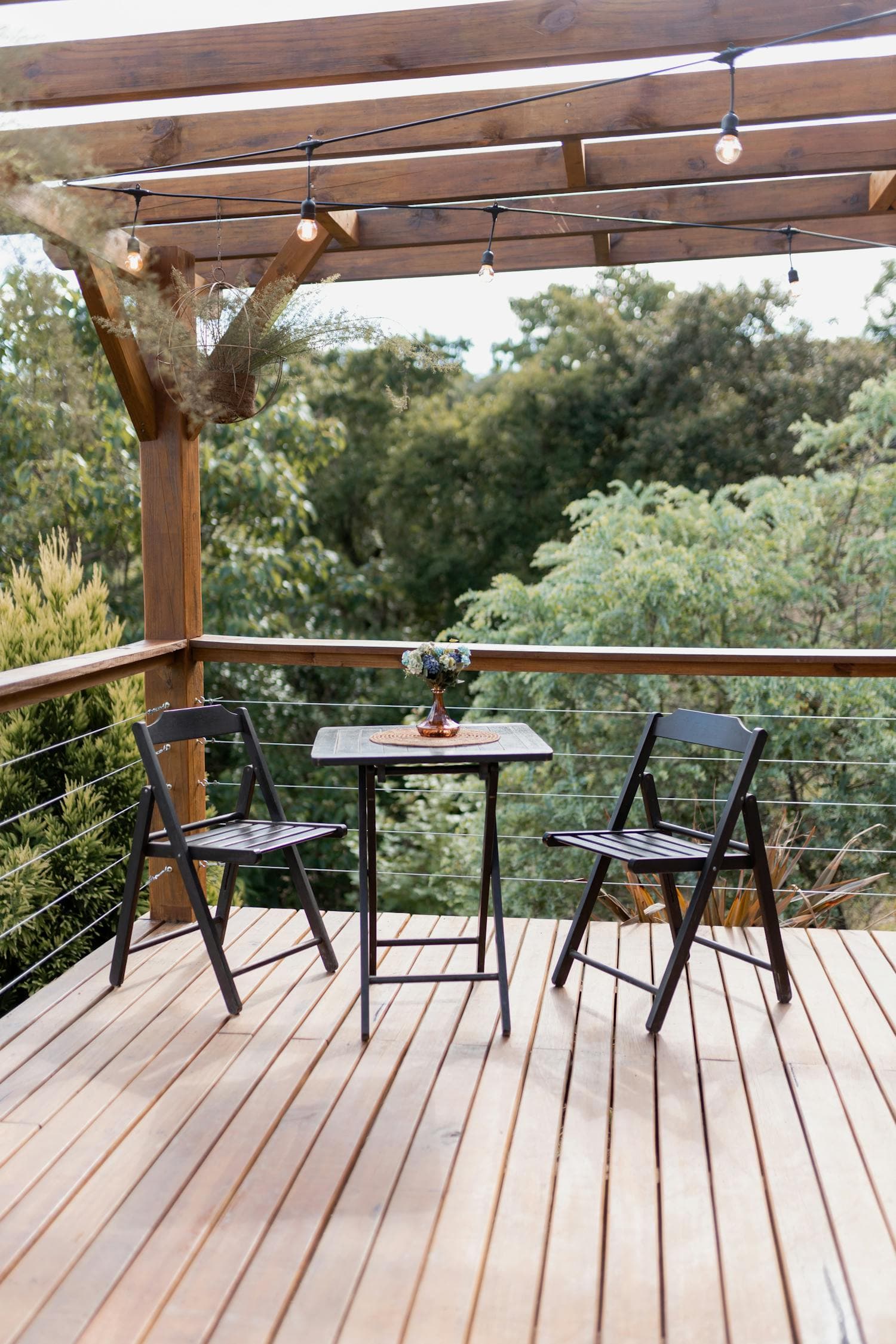A wooden patio with two black folding chairs and a small bistro table under a pergola wrapped in Edison-bulb string lights, overlooking trees in the distance