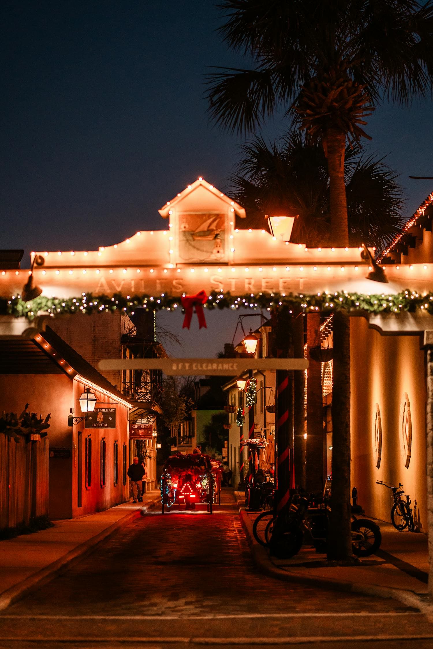 A St. Augustine, Florida side street at twilight with the Aviles Street arch wrapped in warm-white Christmas lights, a large red bow on the garland, and a palm tree visible above the buildings