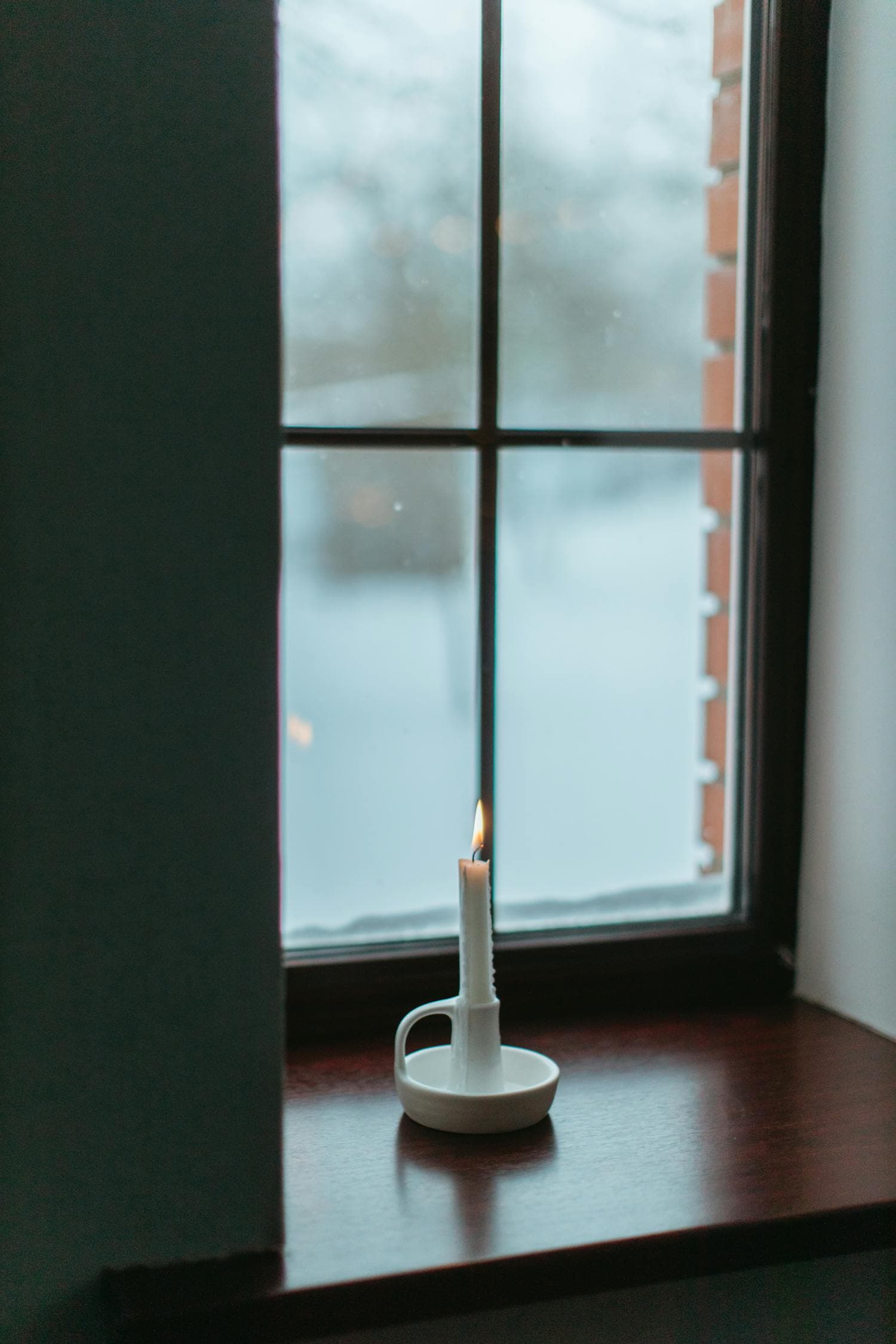 A single warm candle glowing in a window at night with snow visible through the glass outside, the classic colonial-style window-candle move that translates beautifully to a ranch's long horizontal window banks