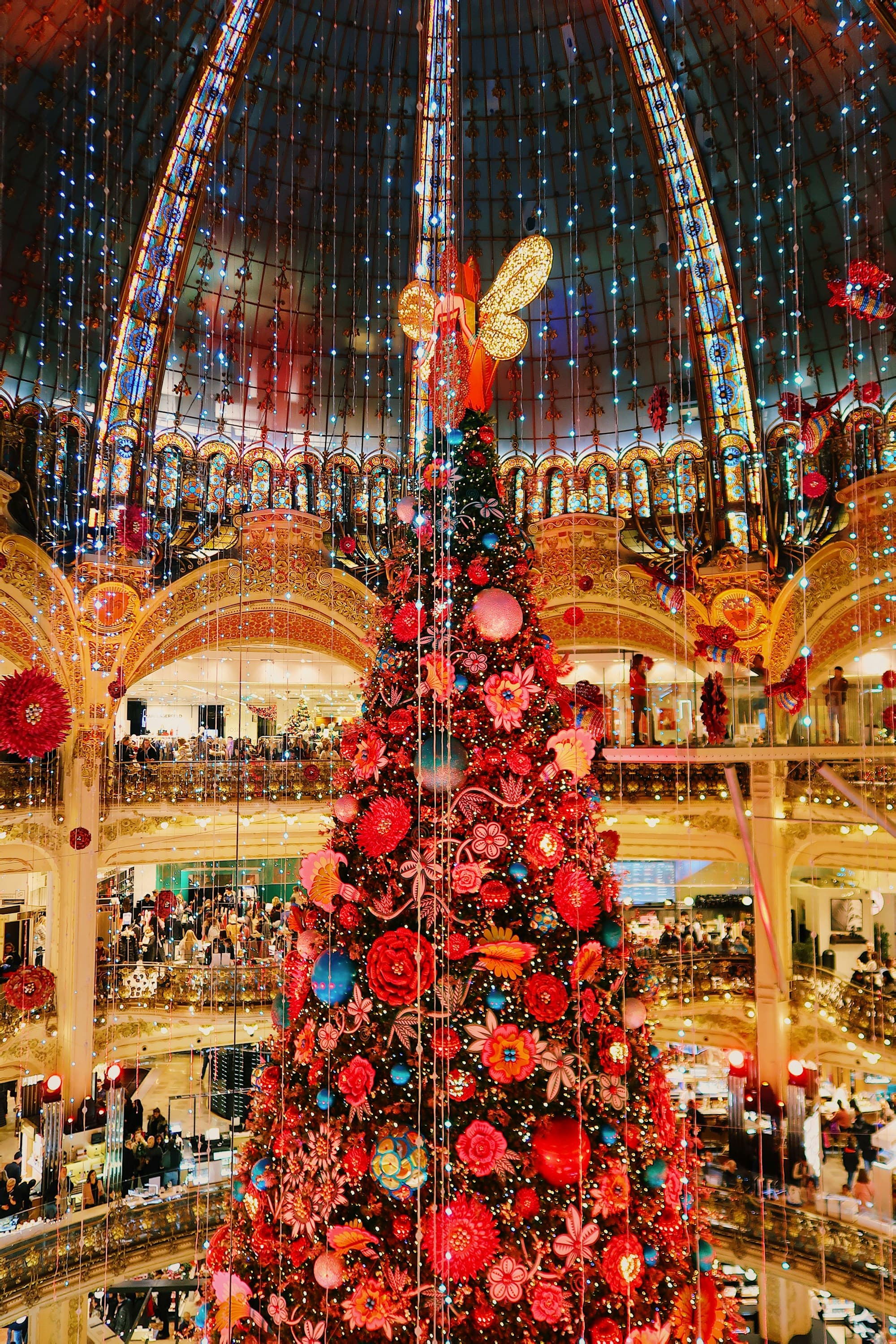 A towering red-and-gold ornate Christmas tree decorated with floral and butterfly motifs standing under the Belle Epoque glass dome of Paris's Galeries Lafayette department store