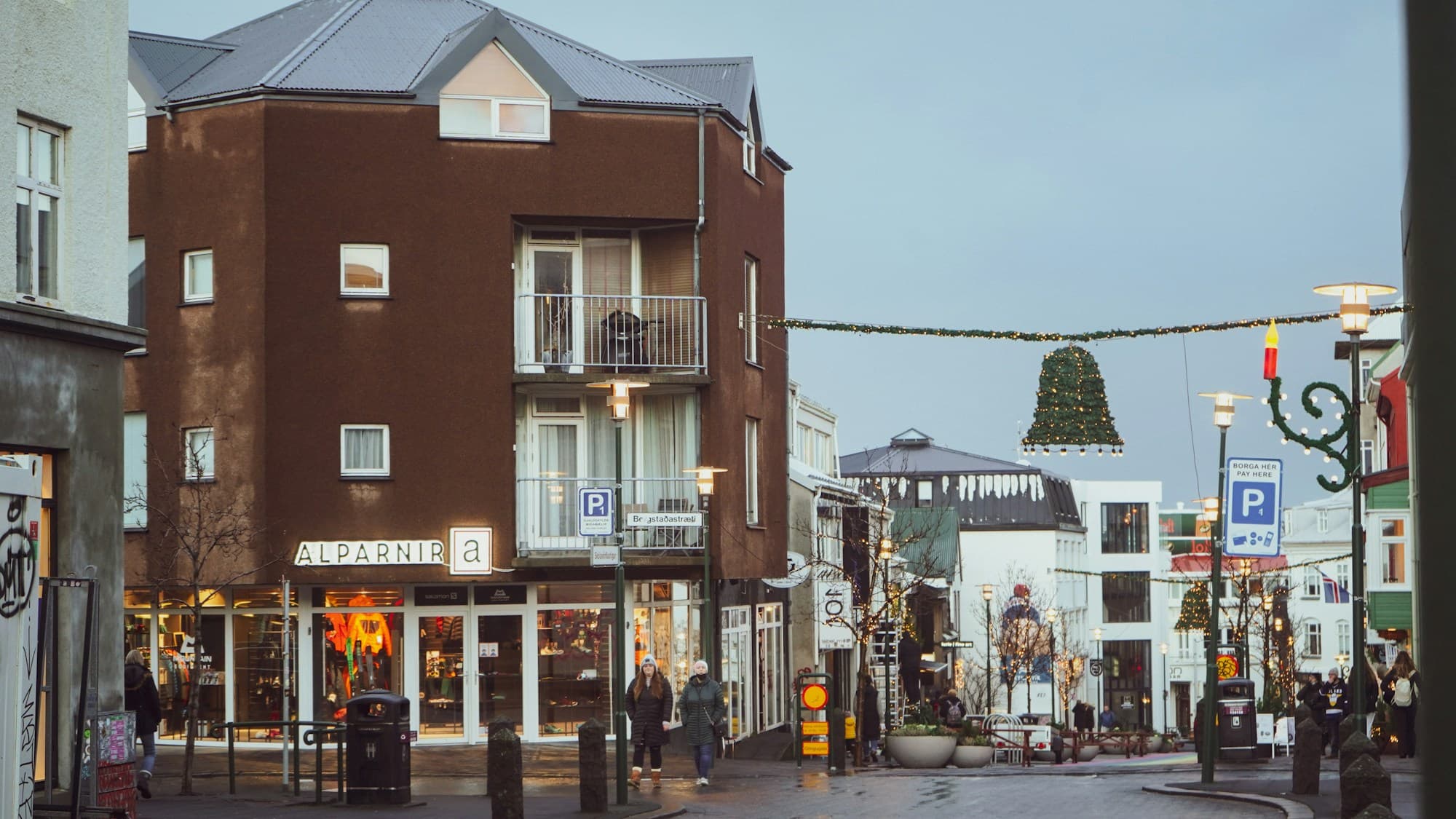 A Reykjavik street at dusk with festive lights strung overhead and a small Christmas tree hanging in the distance, brown wooden buildings and shopfronts lining the cobblestone street
