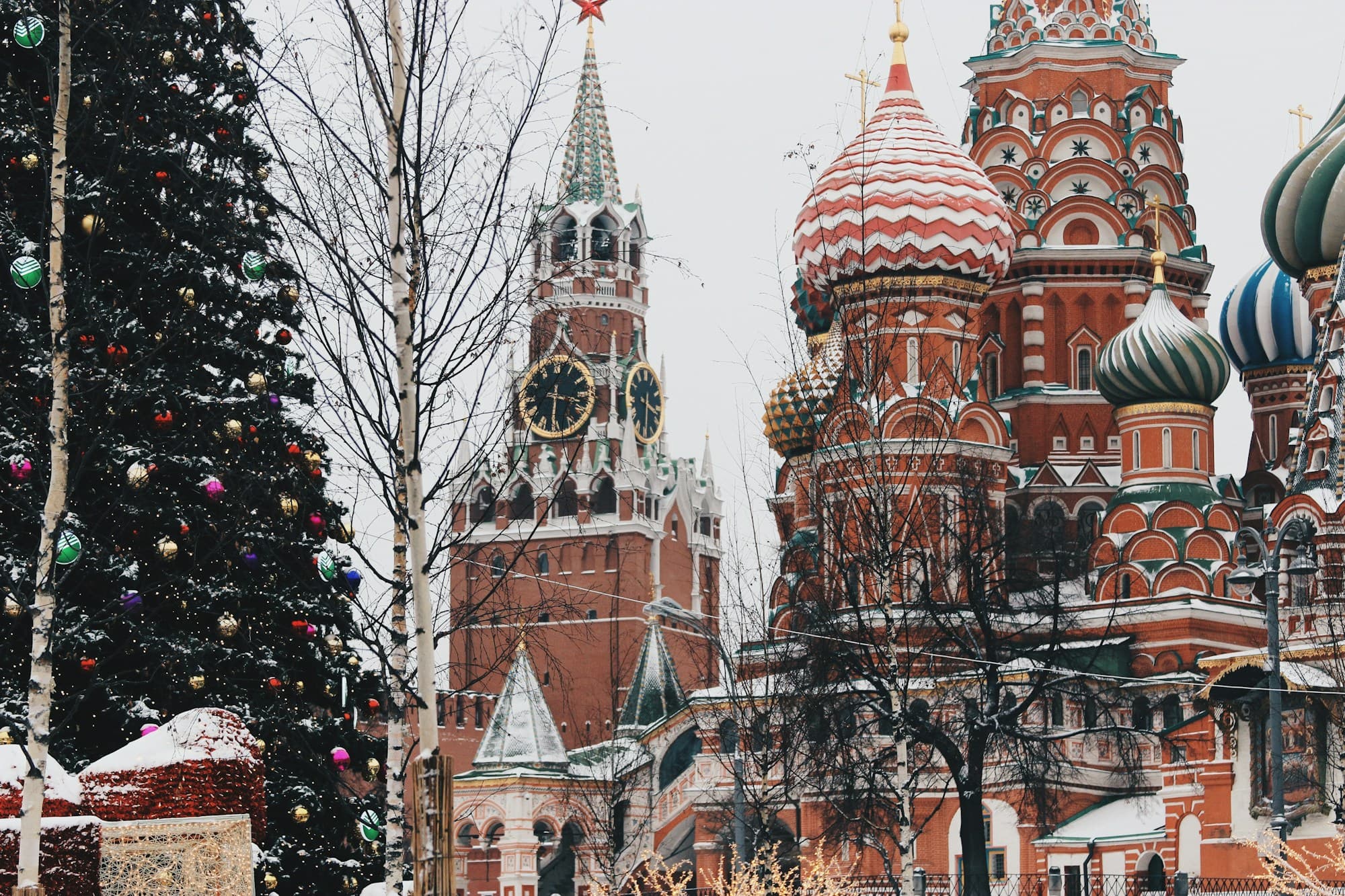 A snow-dusted Moscow Christmas tree decorated with gold and red baubles stands in Red Square next to the Spasskaya Tower's illuminated clock face and Soviet-era red star, with the colorful onion domes of St. Basil's Cathedral rising on the right