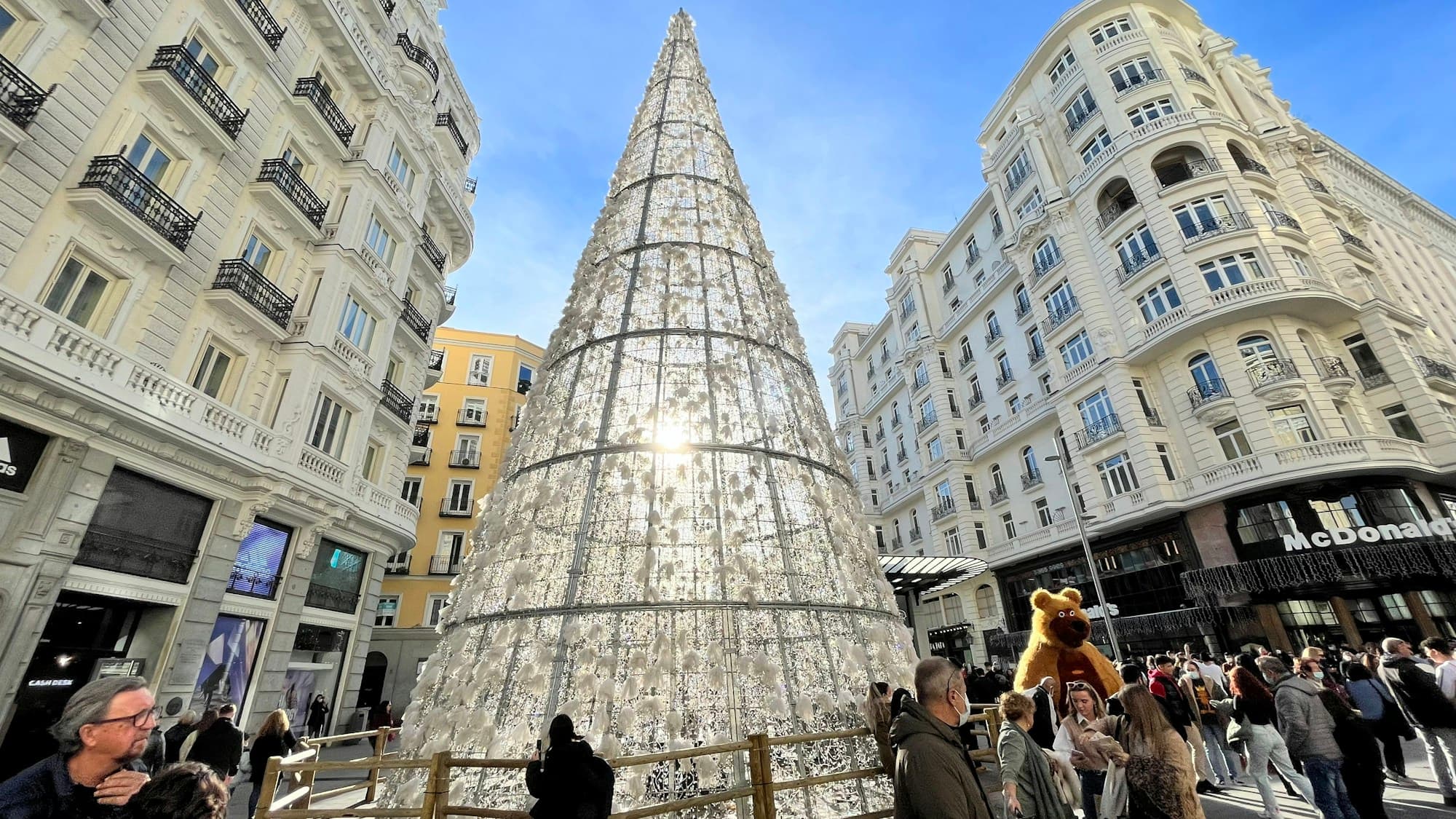 A giant illuminated cone-shaped Christmas tree on Madrid's Gran Via plaza, flanked by classic light-colored Spanish belle-epoque buildings, crowds of bundled visitors gathered around the tree under a blue sky