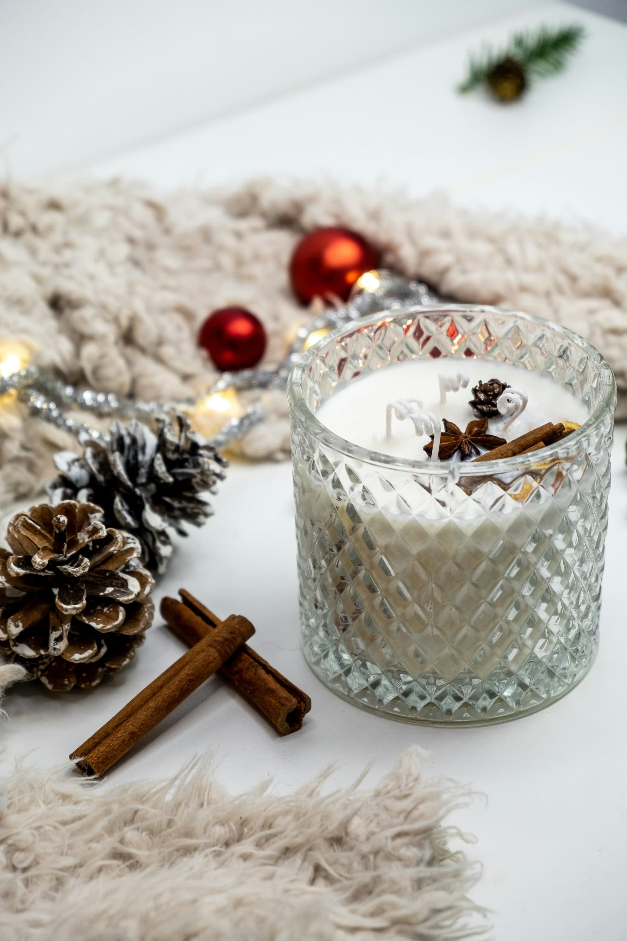 A faceted crystal candle holder with a white candle, surrounded by frosted pinecones, cinnamon sticks, a string of warm white fairy lights, red glass ornaments, and a chunky cream knitted throw - a quintessentially Scandinavian Christmas still life