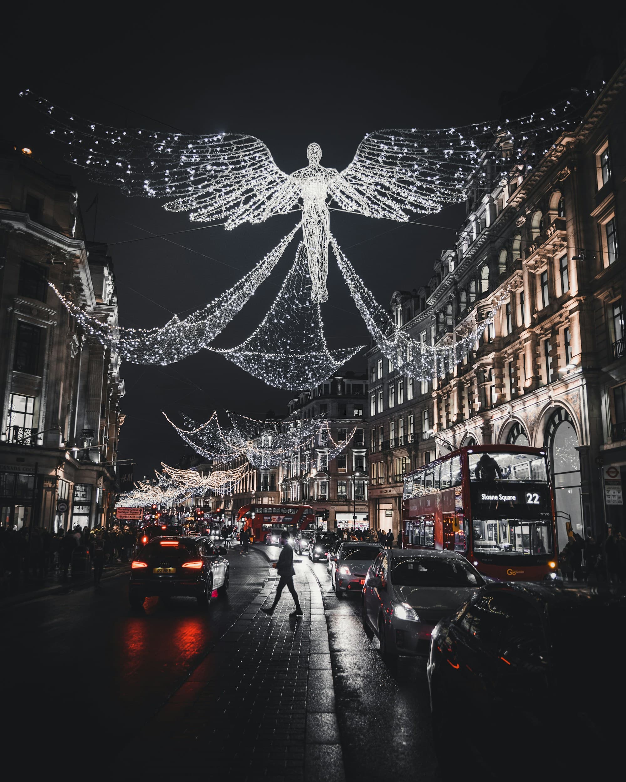 London's Regent Street at night strung with the iconic illuminated angel light artworks suspended above the street, red double-decker buses and a wet pavement reflecting the glow, a pedestrian crossing the street below the angels