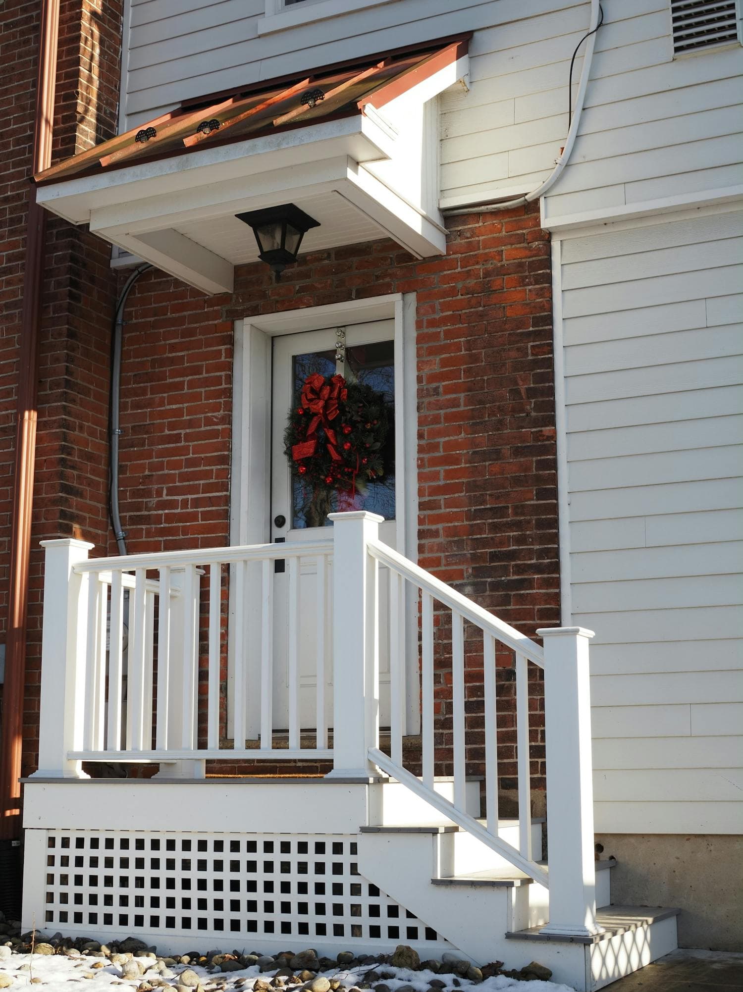 A traditional front porch with stained wood trim and warm-toned siding, decorated with a single fresh evergreen wreath bound with a deep red ribbon hung centered on the door, late-afternoon sunlight slanting across the wood floor