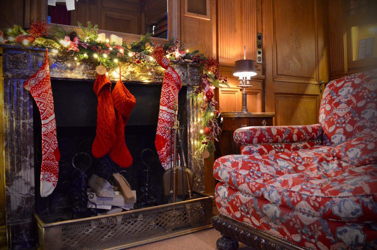 A cozy Craftsman-style living room with a stone fireplace dressed in a fresh evergreen garland, three wool stockings hung from the mantel with copper hooks, and a leather wing chair pulled close to the hearth, all in warm earth tones