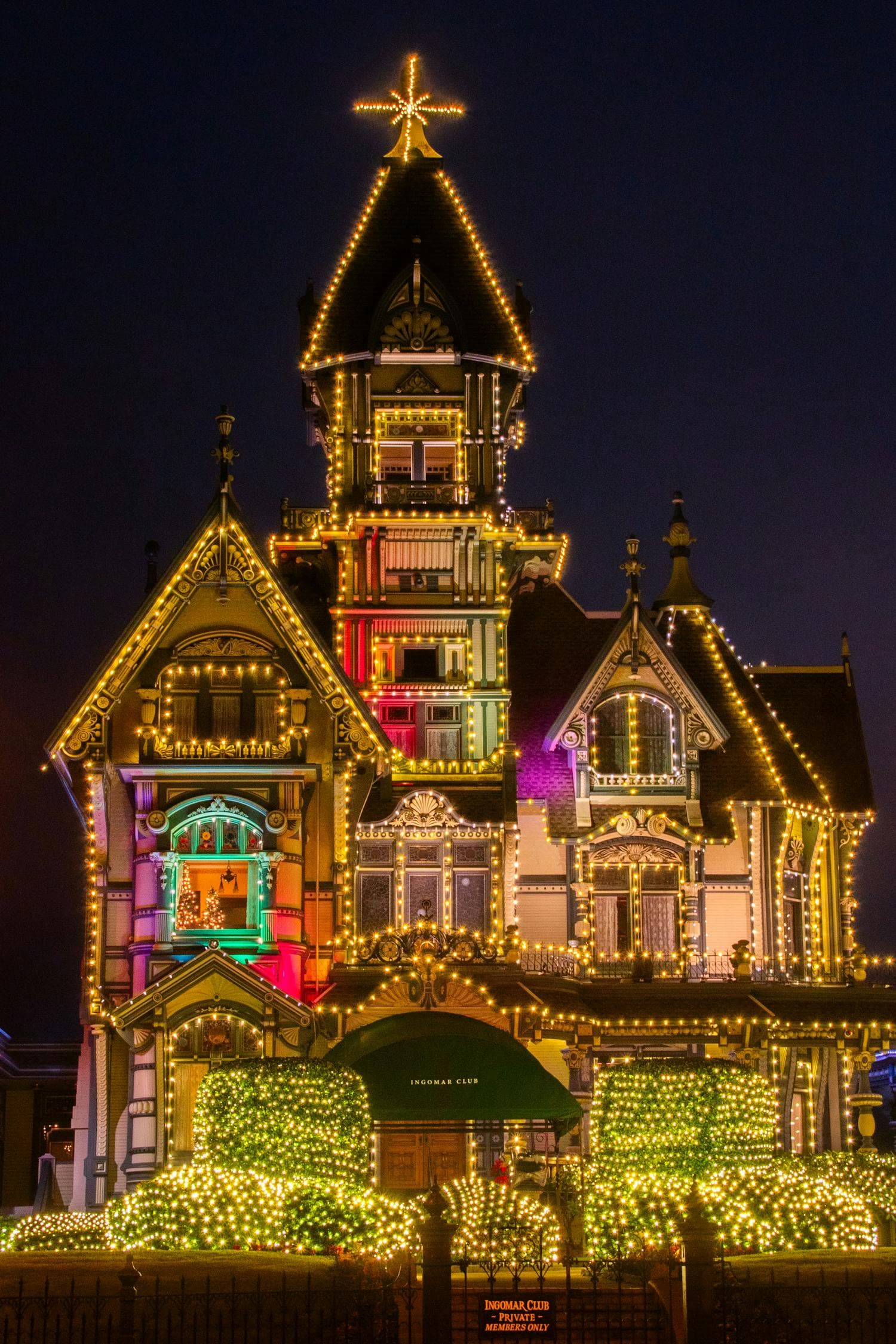 Eureka, California's historic Carson Mansion at twilight, with every architectural feature (turret, gables, spindlework, dormers, and lit hedges in front) outlined in warm-white Christmas lights