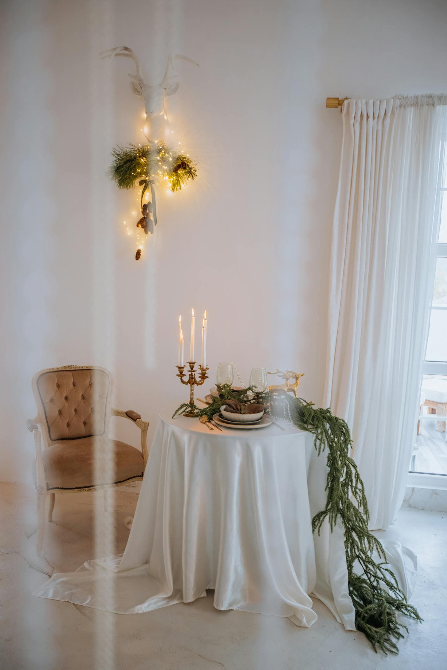 An intimate Victorian-style dining setup with a tufted velvet armchair, a small round table draped in white silk, a brass candelabra holding three lit tapers, a trailing evergreen runner, and a white-painted stag-head wall mount wrapped in fairy lights