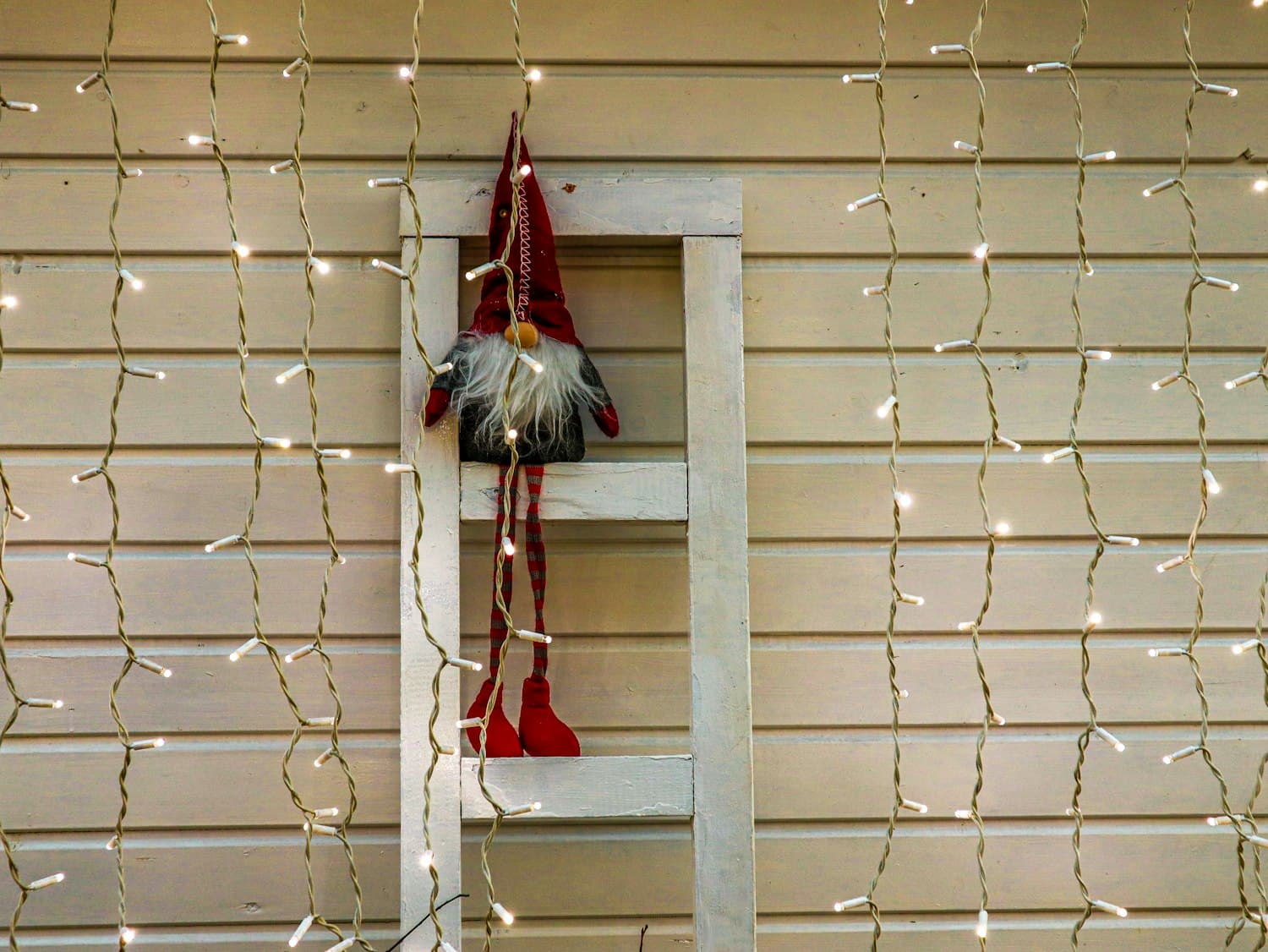 A wooden leaning ladder against beige siding, with cascading warm-white curtain string lights behind it and a small Scandinavian gnome figurine perched on one of the rungs