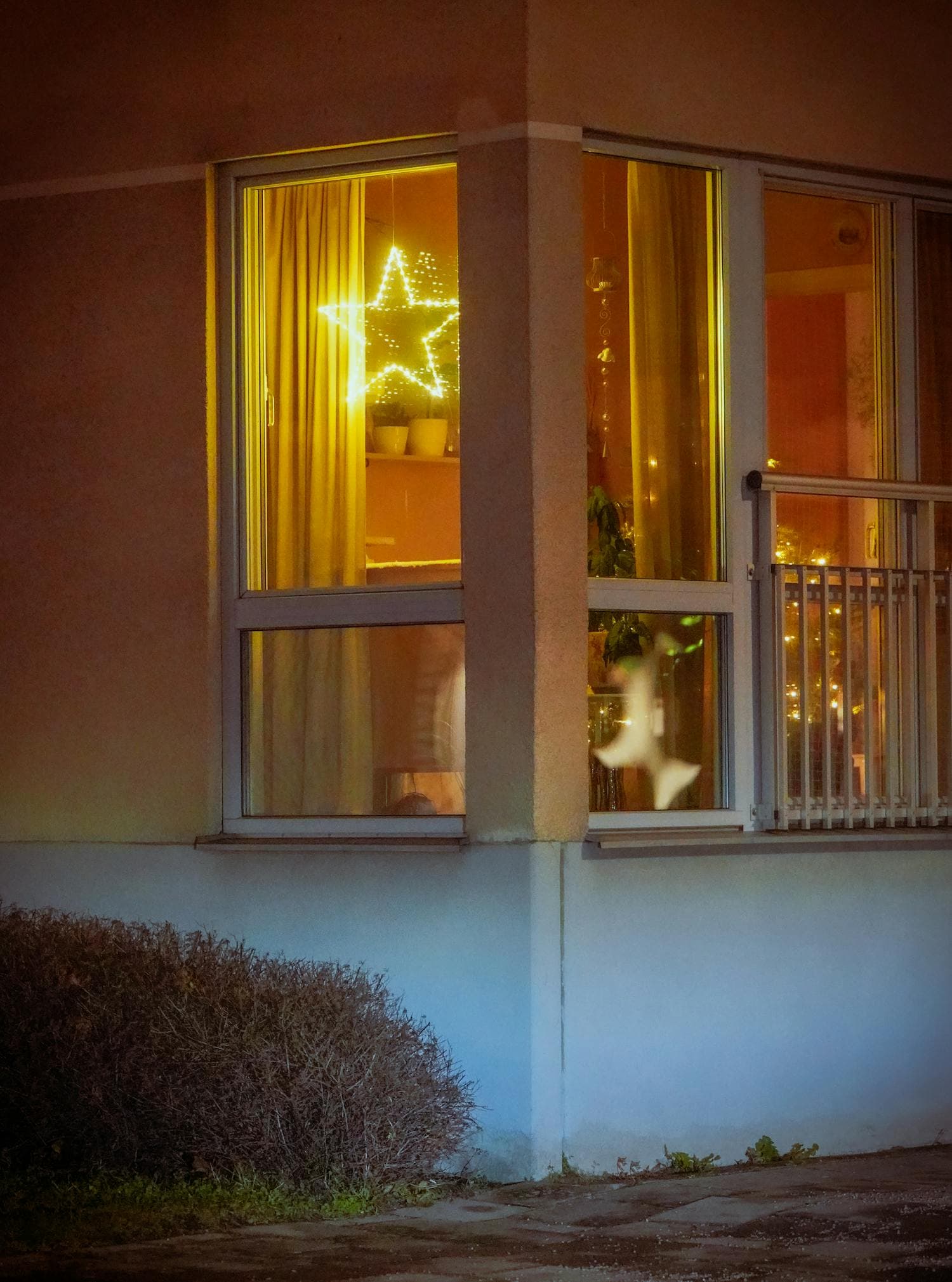 A warm-white star-shaped Christmas light glowing in an apartment window at dusk, the soft golden light catching on the window frame and casting an inviting amber glow over the dark city beyond