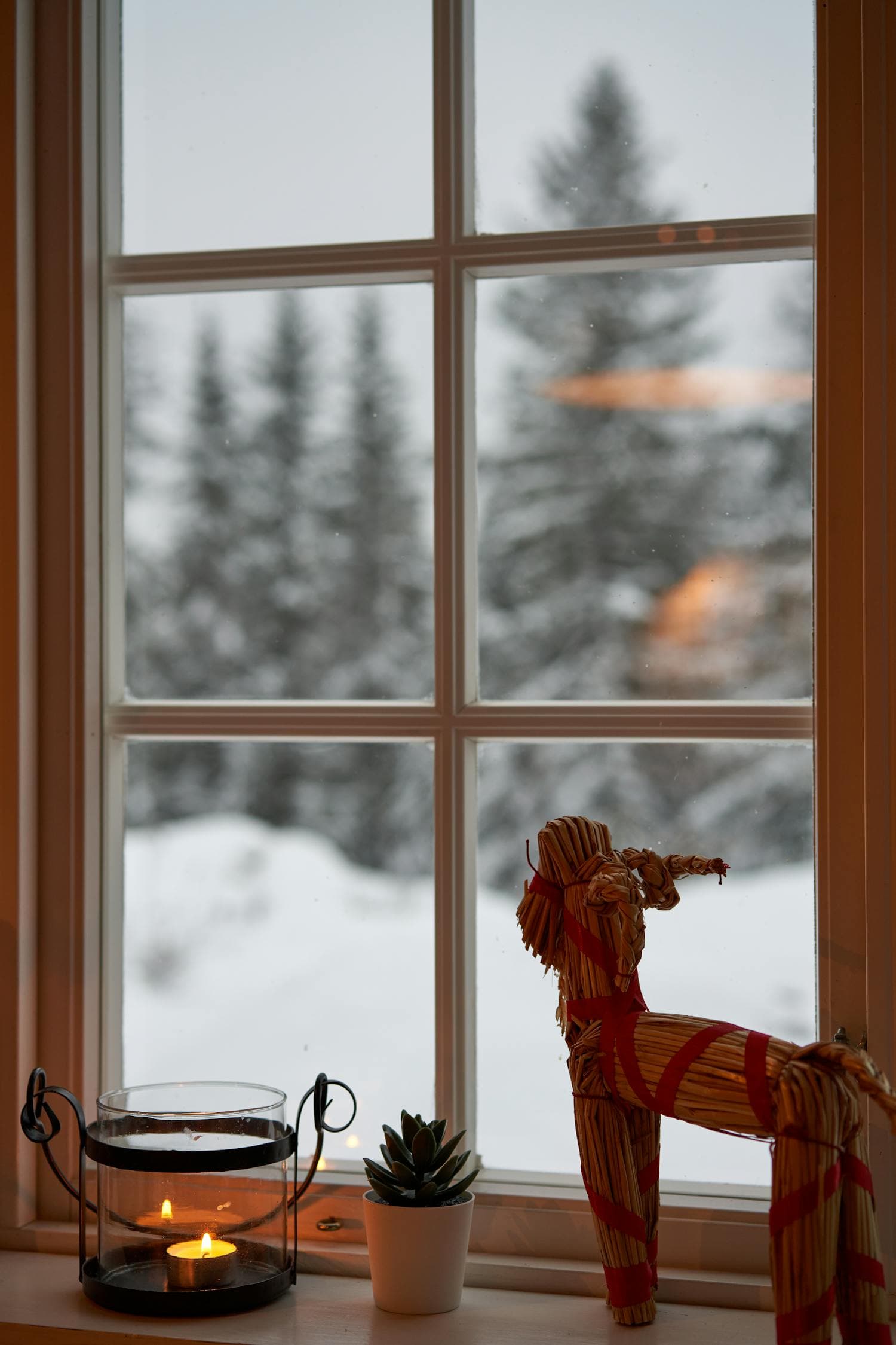 A windowsill at twilight with a single lit candle burning warmly beside a small wooden Yule goat (julbock) figurine, snow visible through the window, the kind of restrained Scandinavian Christmas vignette built around atmosphere rather than ornament