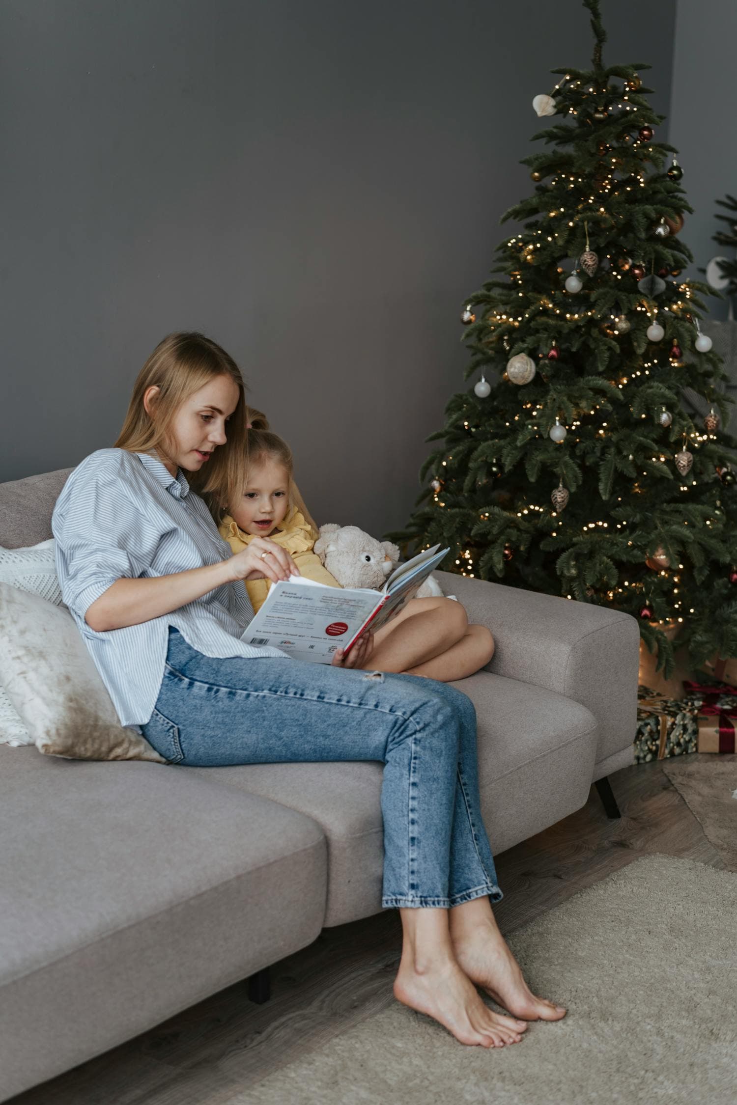 A mother and daughter snuggled together on a gray couch reading a book, with a softly lit decorated Christmas tree in the background, the quintessential cozy December reading moment