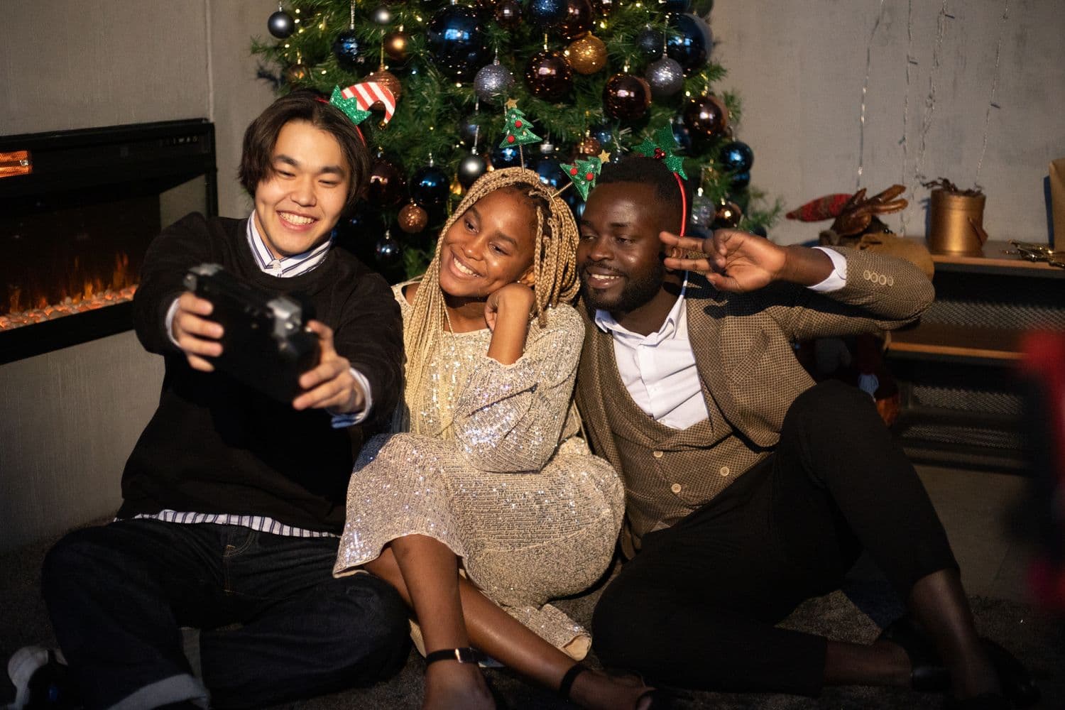 Three friends in festive clothes sitting together by a decorated Christmas tree and fireplace, laughing and taking a selfie at a holiday gathering