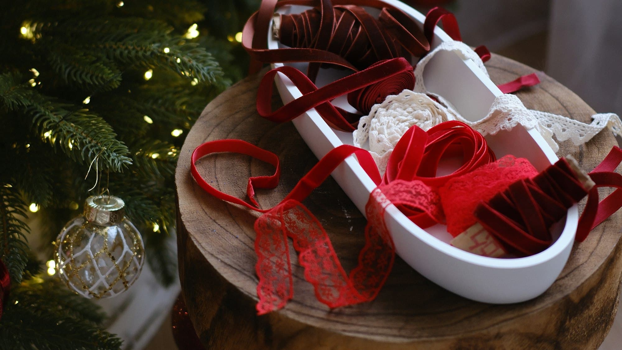 Spools of red velvet and lace Christmas ribbon in a dish on a wooden stool beside a lit tree