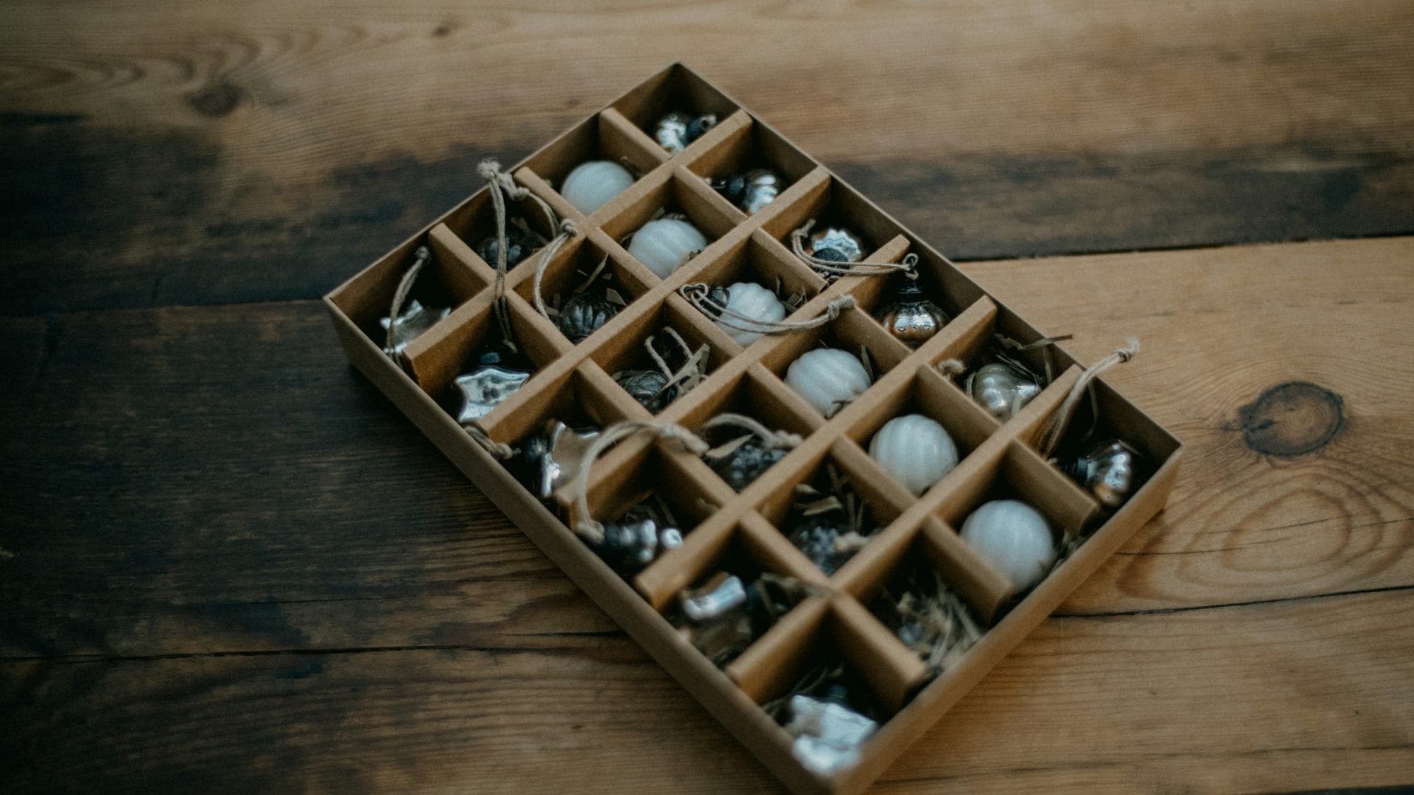 A divided cardboard ornament storage box on a wood floor, each compartment holding a white or mercury-glass Christmas ornament
