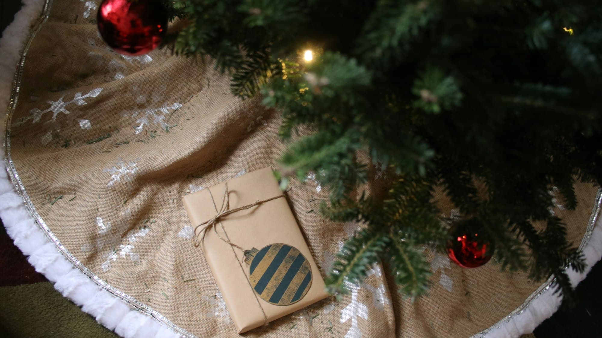 A burlap Christmas tree skirt with a fur-trimmed edge under a decorated tree, with a kraft-wrapped gift on top
