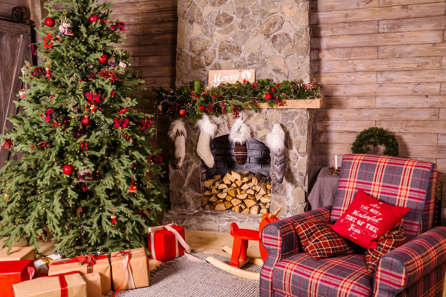Traditional Christmas living room with a decorated tree in red ornaments, presents underneath, stone fireplace with stockings and a garlanded mantel, and a red-and-green plaid armchair
