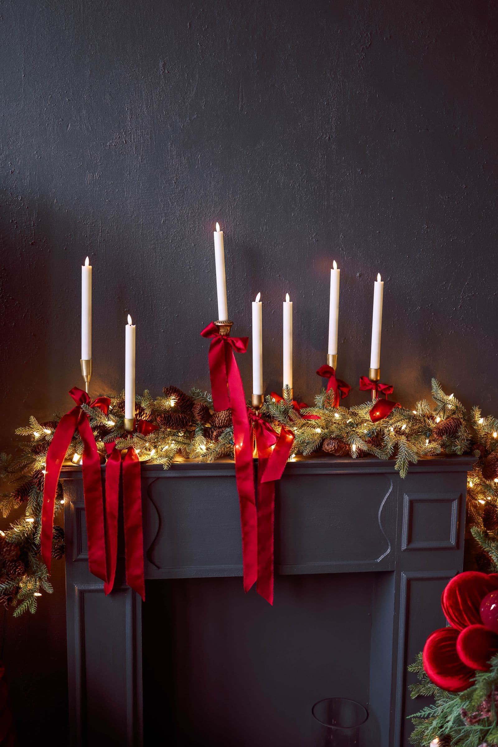 White taper candles in brass holders with trailing red velvet ribbons on a dark mantel dressed in lit pinecone garland