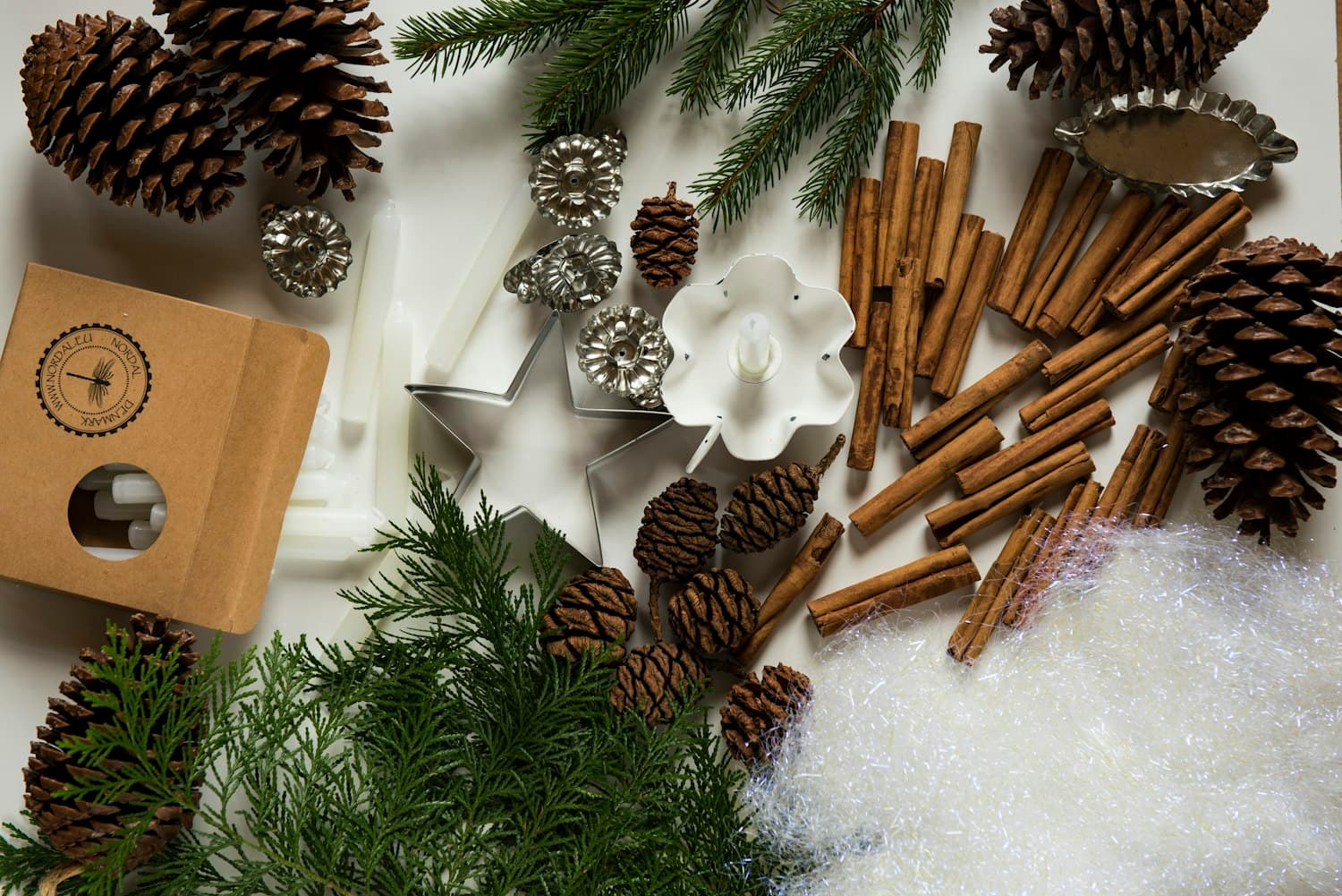 Overhead flat lay of Christmas planning supplies, pinecones, cinnamon sticks, white candles, evergreen sprigs, star cookie cutter, tinsel, kraft-paper box
