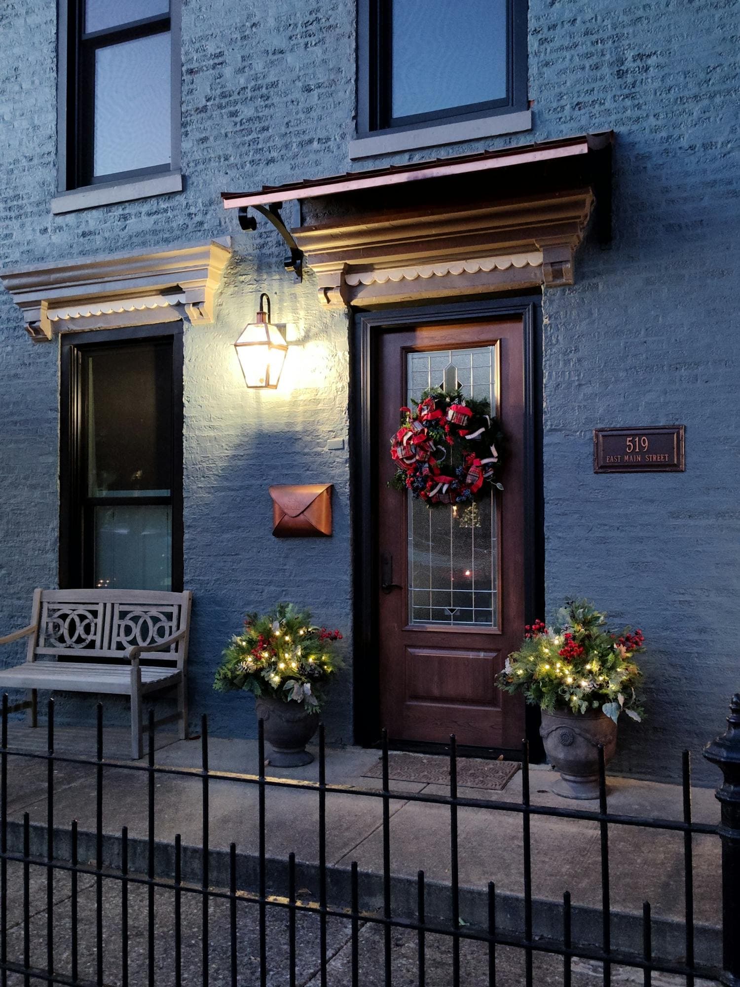 A covered front entry at twilight with a Christmas wreath on the door, a lit gas lantern overhead, and matched evergreen planters with string lights flanking the steps