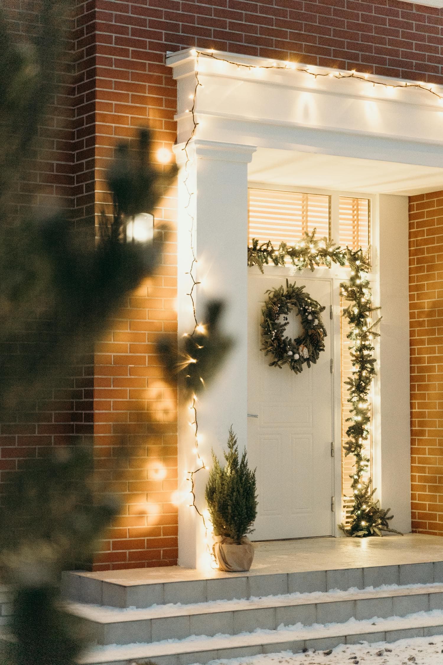 A white-columned brick-home entry with warm-white string lights wrapped from base to capital along the porch column, evergreen garland framing the front door, and a lit wreath on the door, the column-wrapping move that defines a Craftsman porch at Christmas