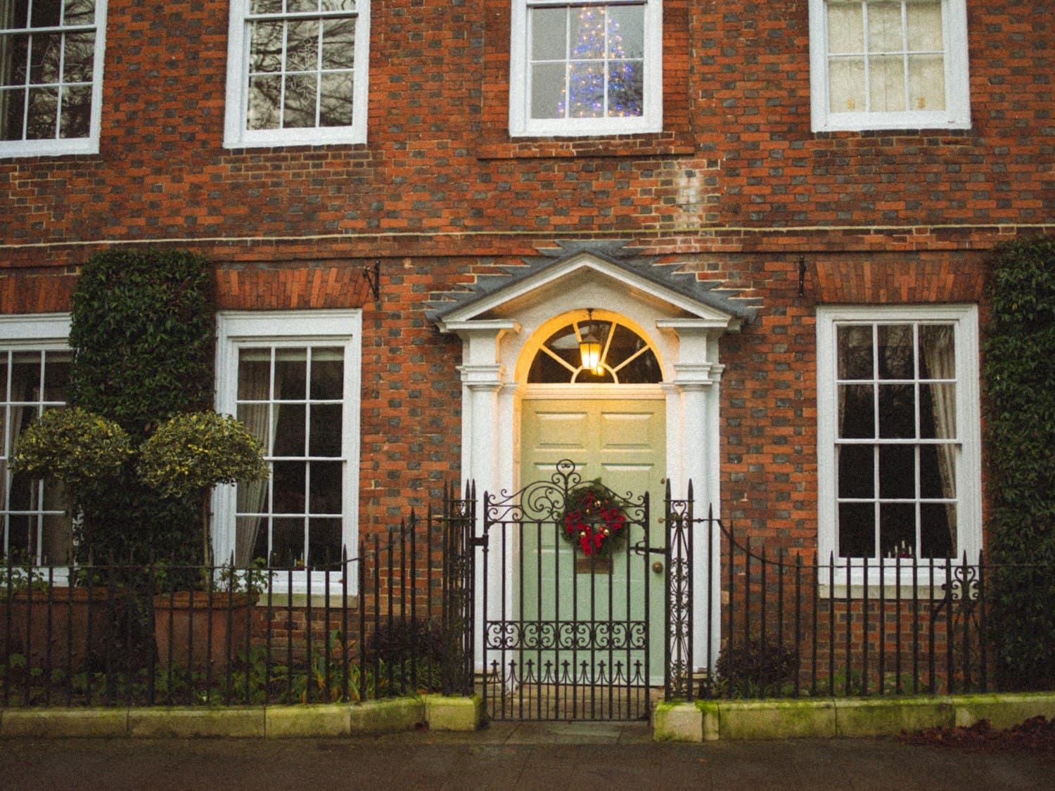 A Georgian brick home with a sage-green front door, classical pediment overhead, and a single Christmas wreath with red ornaments scaled neatly to the door, a textbook example of decor that frames the door instead of burying it, plus a Christmas tree visible through the upstairs window