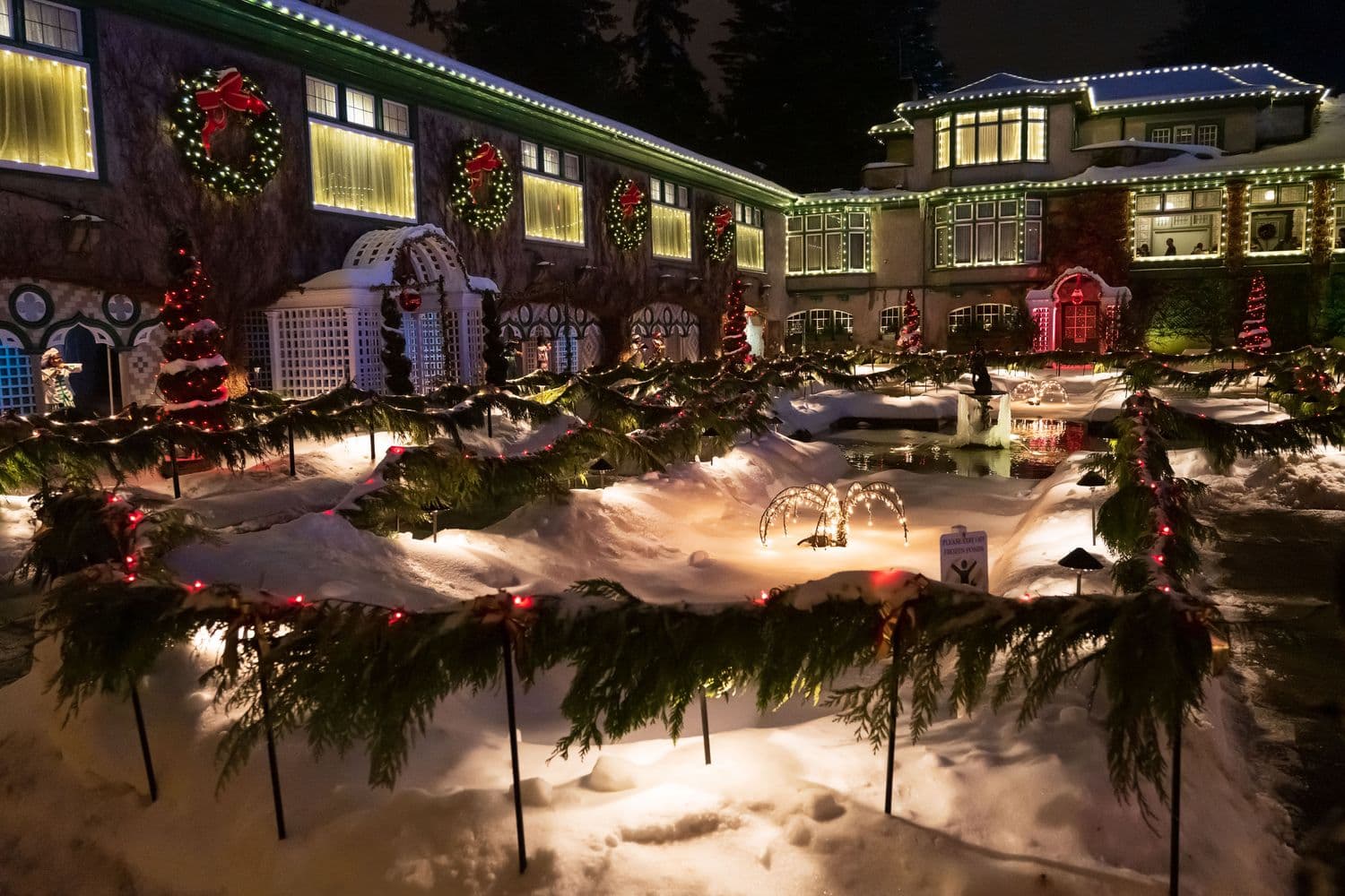 A large estate elaborately decorated for Christmas at night, warm white lights outlining the rooflines, wreaths on the windows, and lit garland lining the snowy garden paths