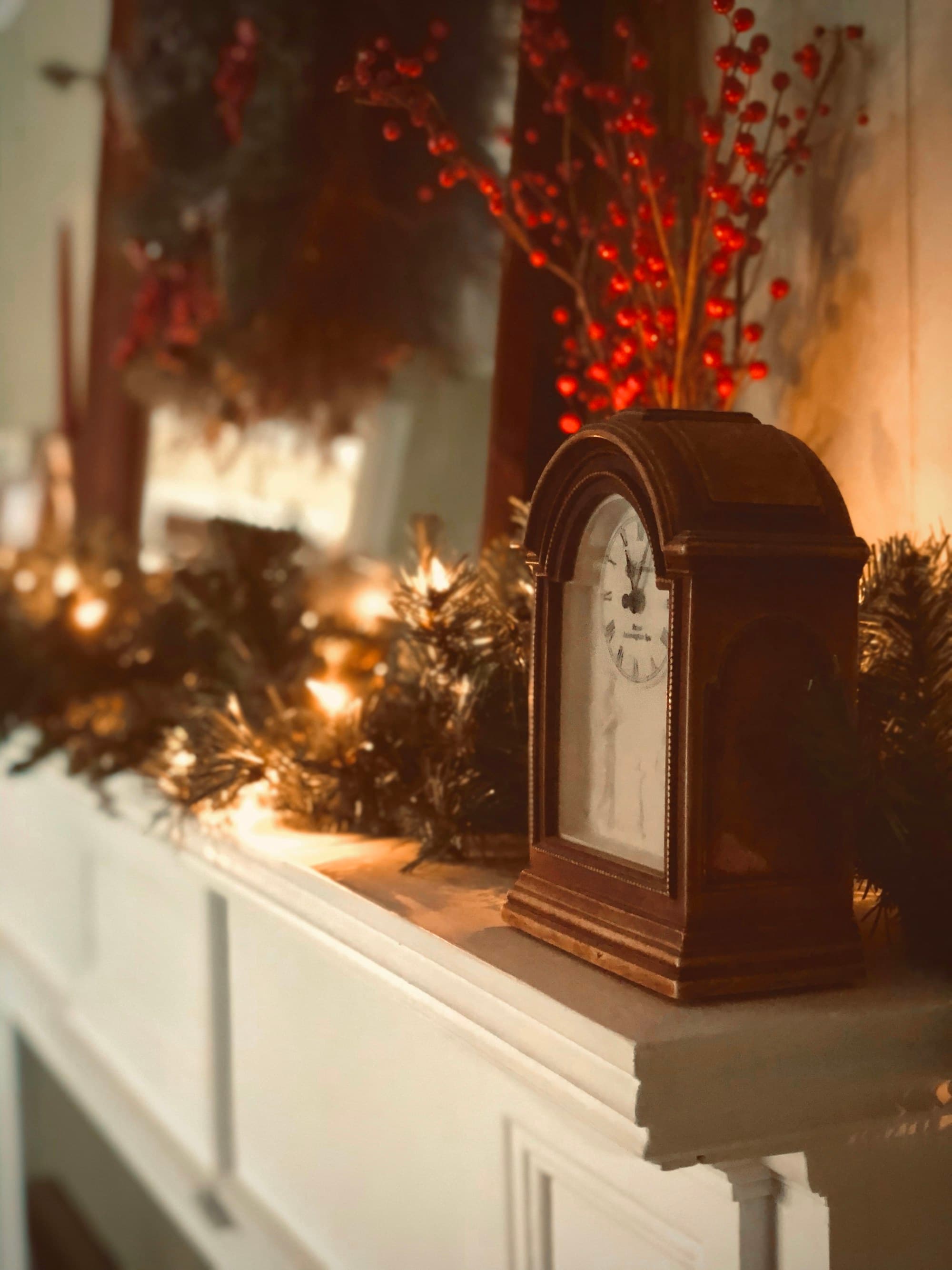 A white painted mantel decorated for Christmas with cedar garland, warm string lights woven through the greenery, and an antique wood mantel clock as the centerpiece, photographed in warm interior dusk light