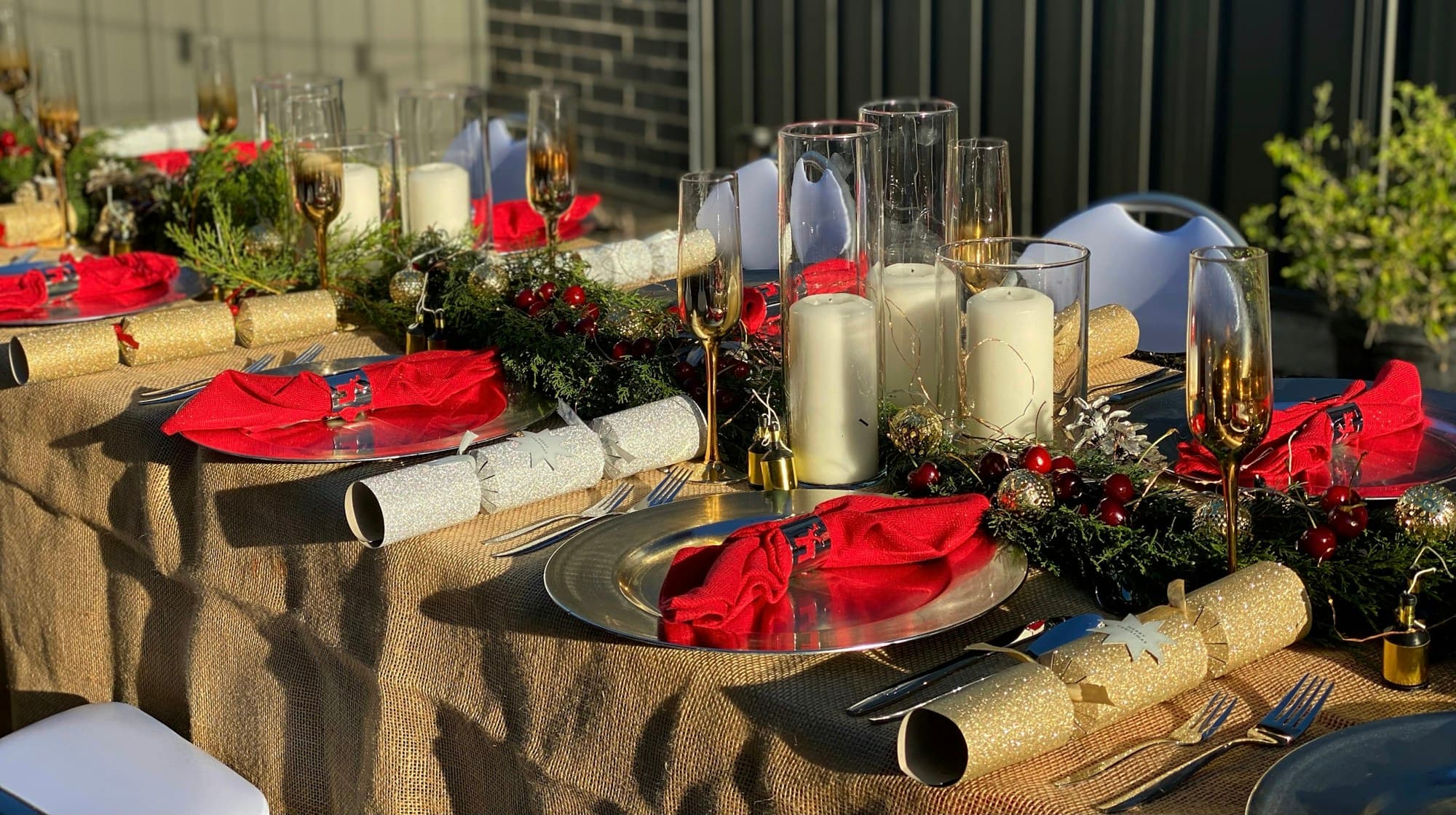 A Christmas table set along its length with a burlap runner, gold chargers, red cloth napkins, and a low centerpiece of fresh cedar greenery studded with red berries and ornaments, white pillar candles and gold champagne flutes catching the warm light
