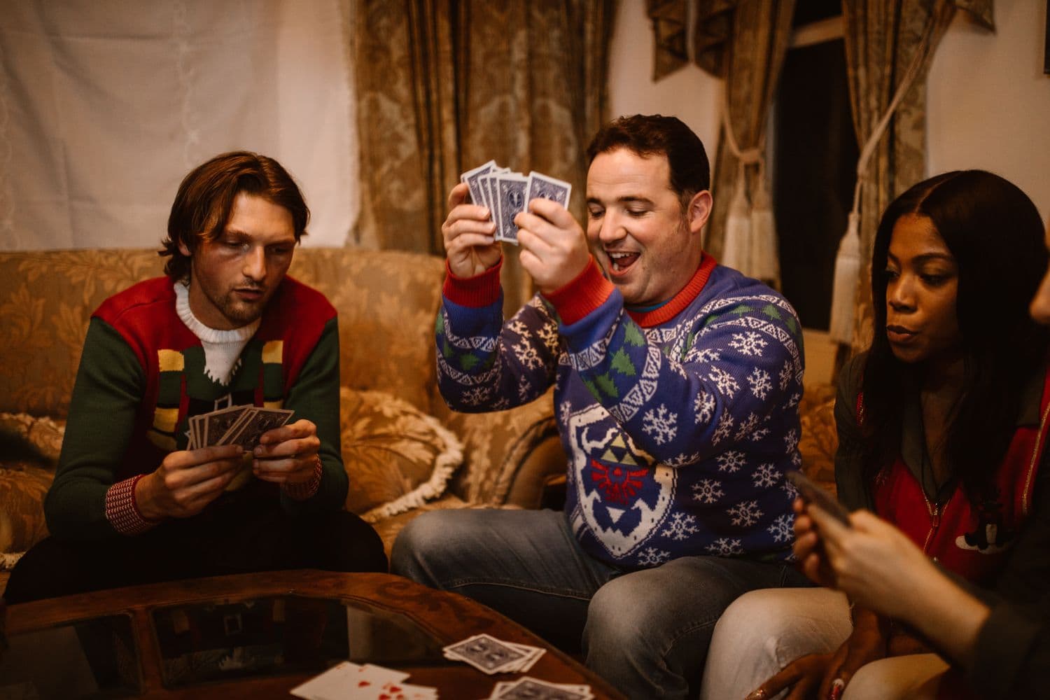 Three friends in festive Christmas sweaters on a couch playing a card game, one laughing as he holds up his hand of cards