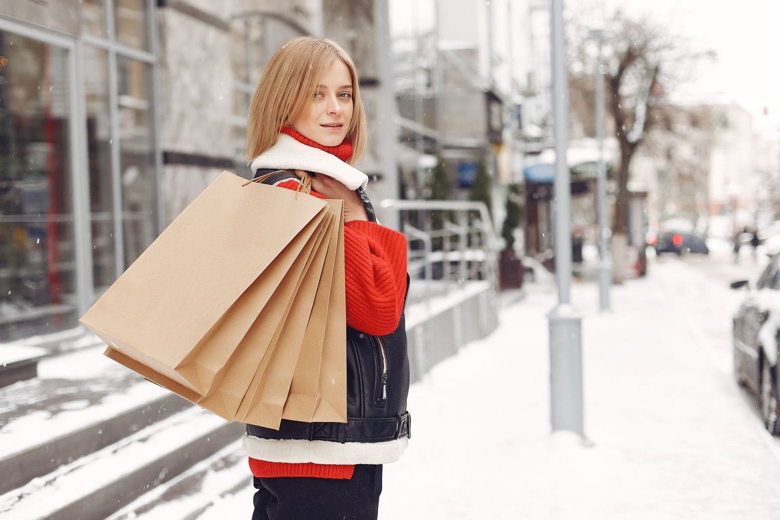 A woman in a red sweater carries an armful of kraft-paper shopping bags along a snowy shopfront-lined street during the holidays