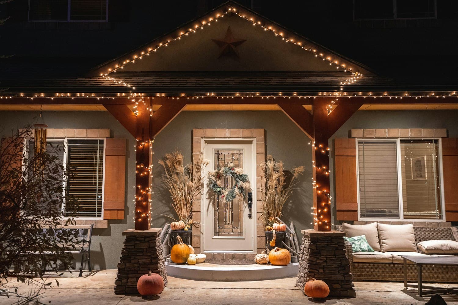 Craftsman-style covered front porch at night with tapered columns on stone bases wrapped in warm white string lights, exposed beam ceiling, wreath on the front door