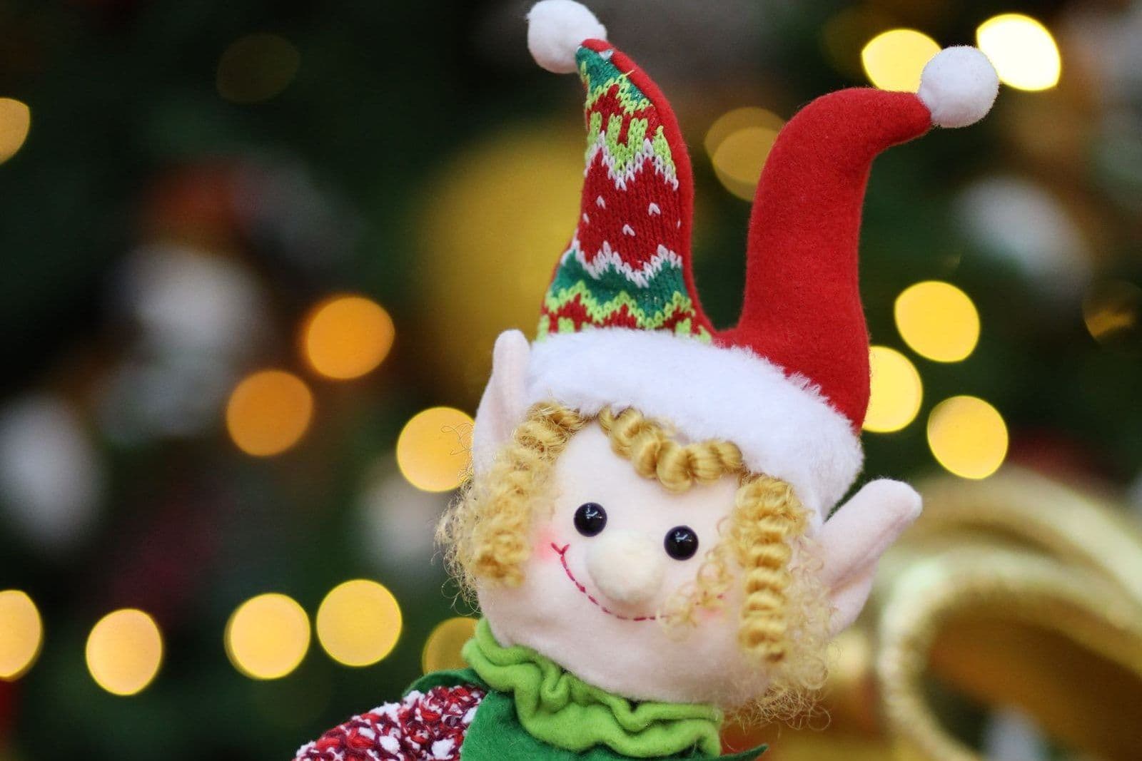 A soft Christmas elf doll with blonde curls and a red and green pointed hat, with warm out-of-focus tree lights behind it