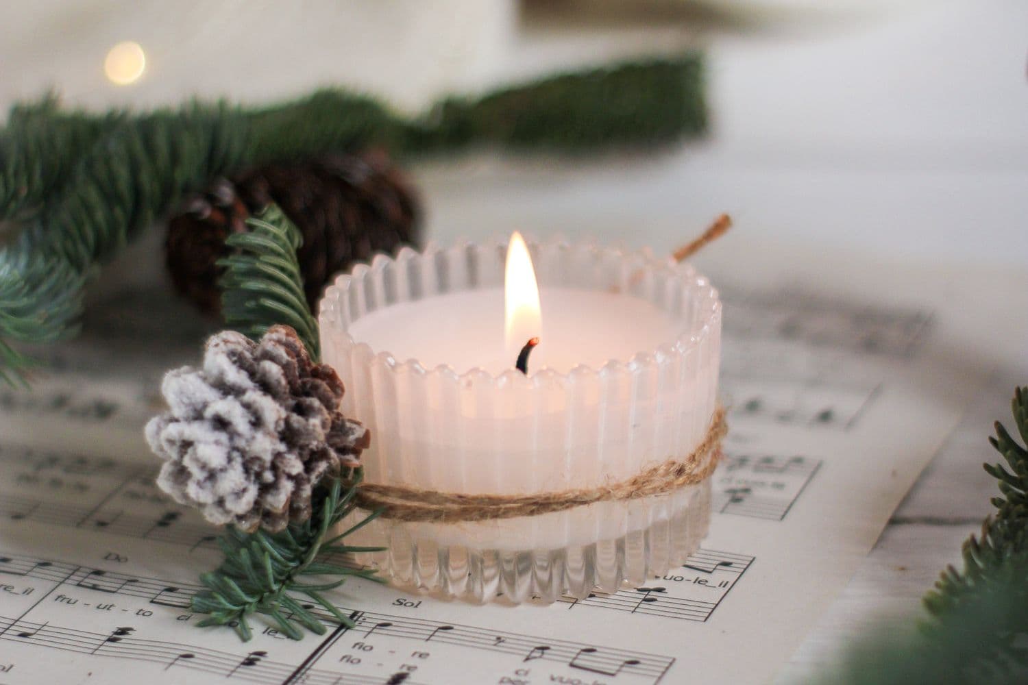 A lit white candle wrapped in twine resting on old sheet music, with pine sprigs and a frosted pinecone alongside