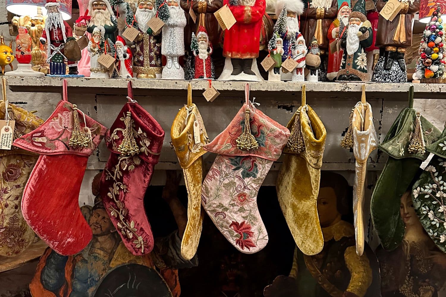 A shelf of vintage Old-World Santa Claus figurines from many eras above a row of antique embroidered Christmas stockings
