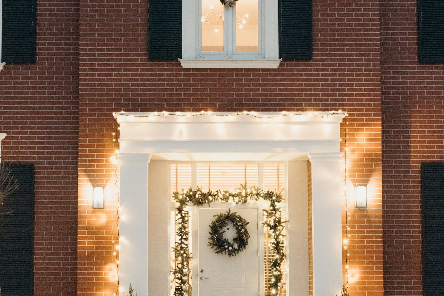 A brick house entry at dusk outlined in warm white string lights, with a lit garland framing the front door and an evergreen wreath