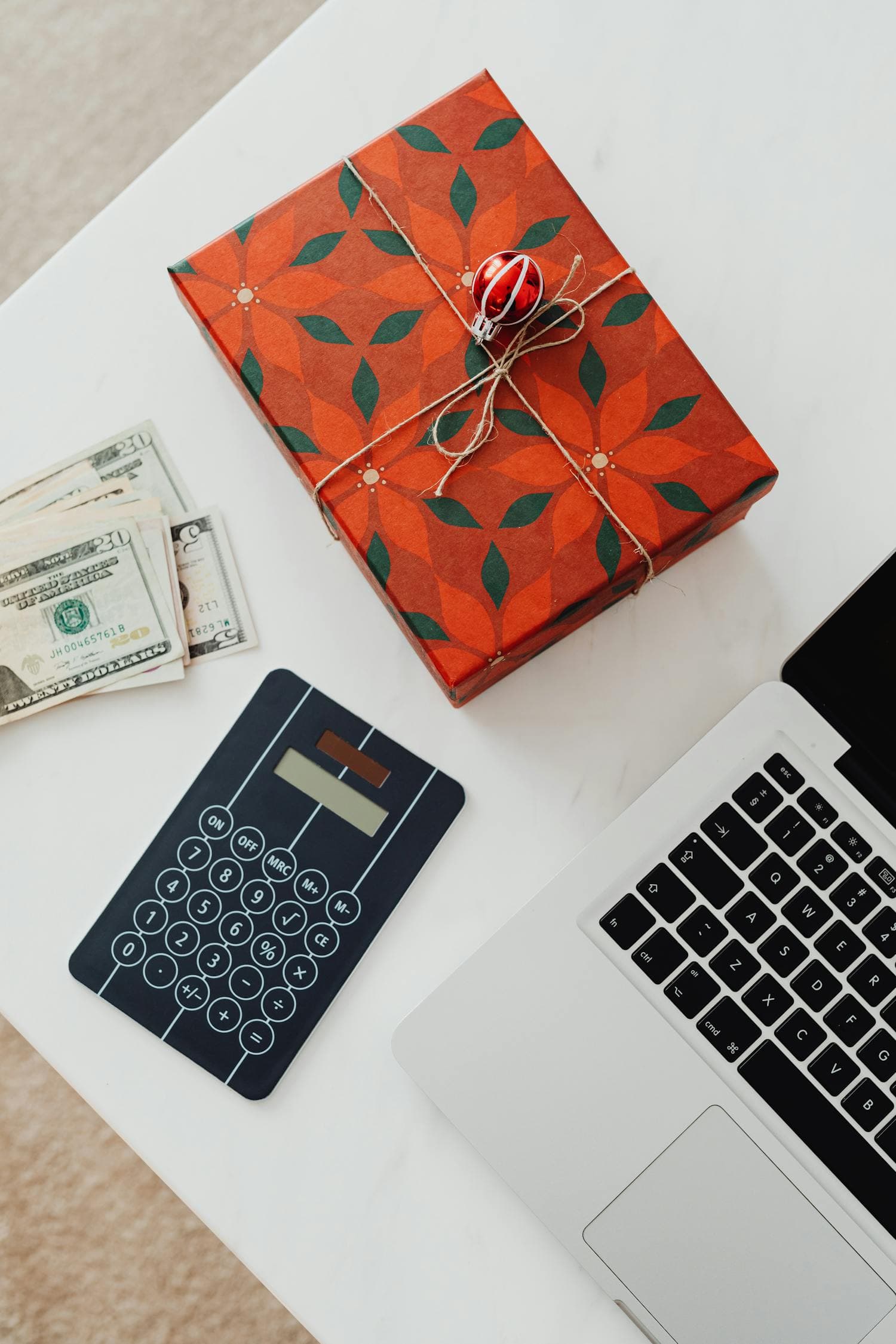 Overhead flat lay of a red poinsettia-print wrapped Christmas gift box with a small ornament tied into the twine bow, beside a stack of $20 bills, a black calculator, and the corner of a silver laptop on a clean white desk