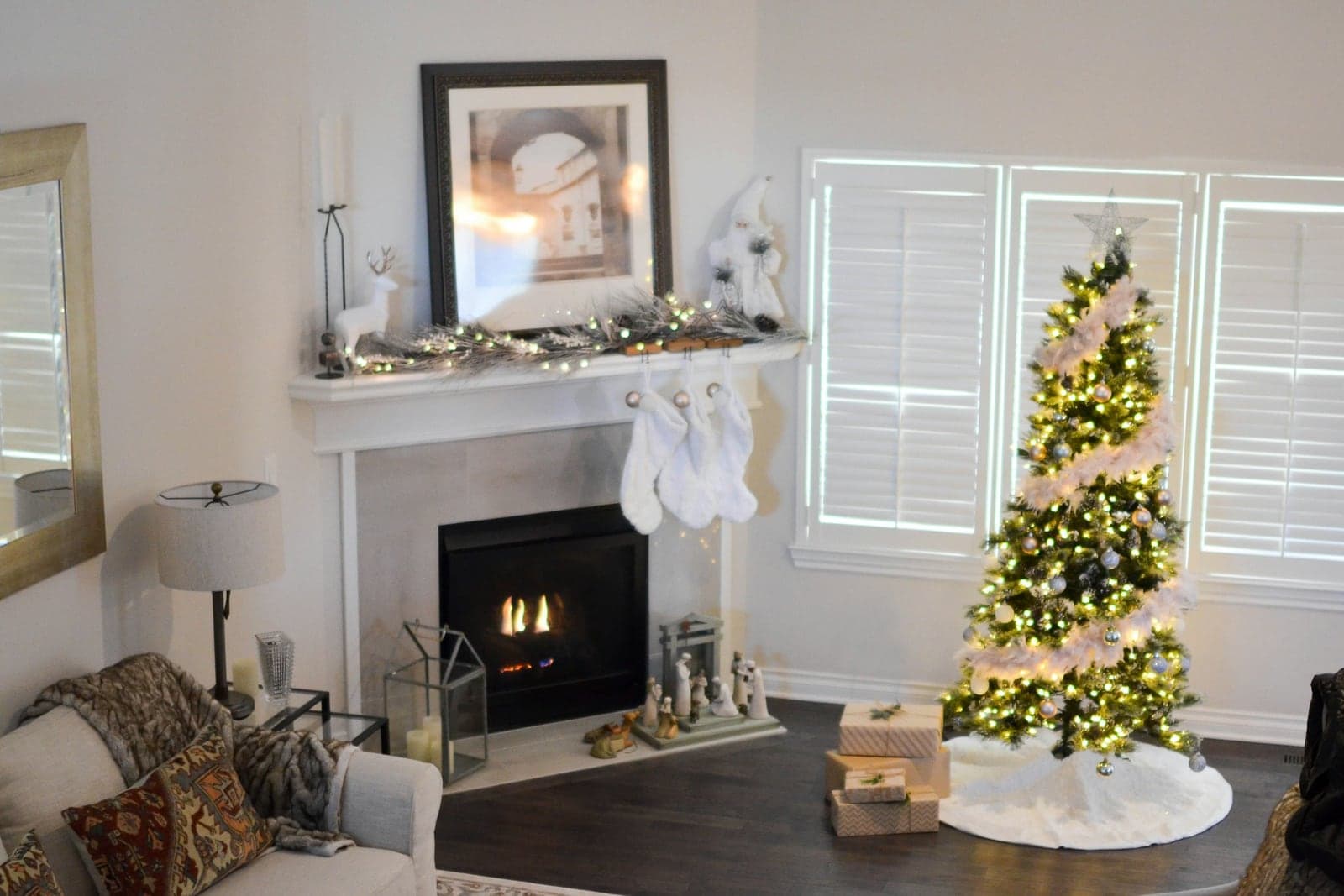 A fully decorated Christmas tree with warm white lights, soft garland, a star topper, and a white tree skirt with wrapped presents, next to a fireplace with stockings