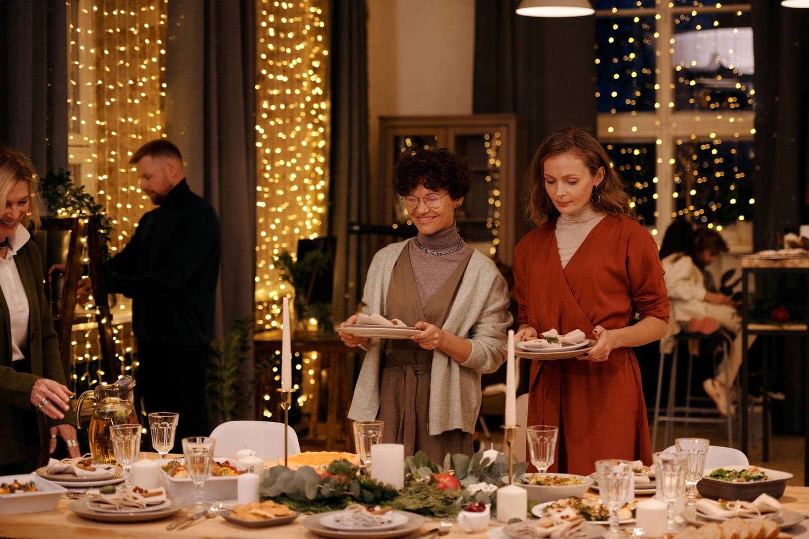 Two women carrying plates to a long candlelit holiday table set with greenery, glassware, and platters, with warm string lights covering the windows behind them