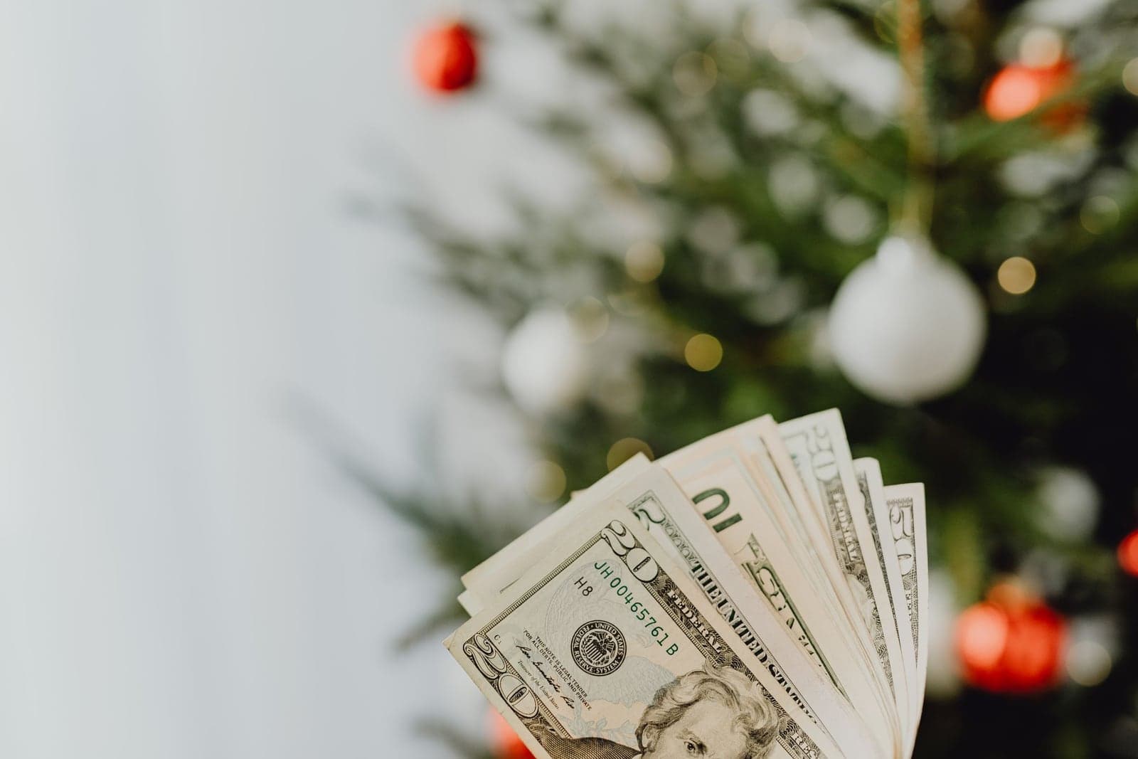 A fan of twenty dollar bills held up in front of a softly blurred Christmas tree decorated with white and red ornaments and warm lights