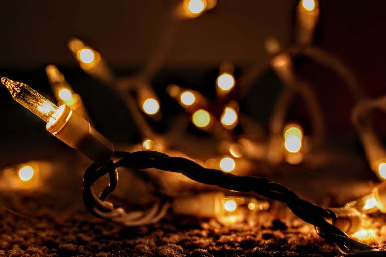 A single warm-glowing mini Christmas bulb in sharp focus with a soft golden bokeh of string lights behind it on a dark background
